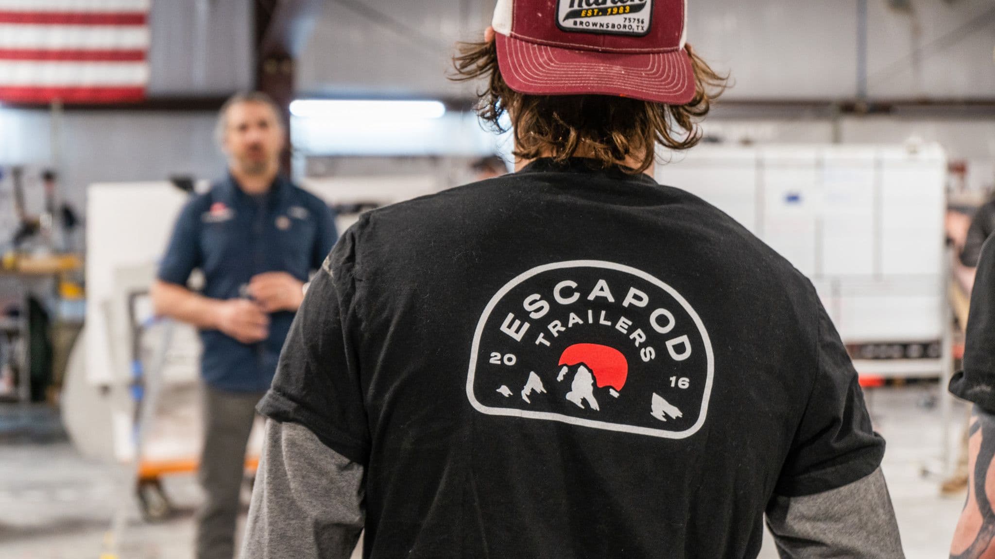 A close-up of a worker wearing an Escapod Trailers T-shirt, standing in the factory with an American flag in the background.