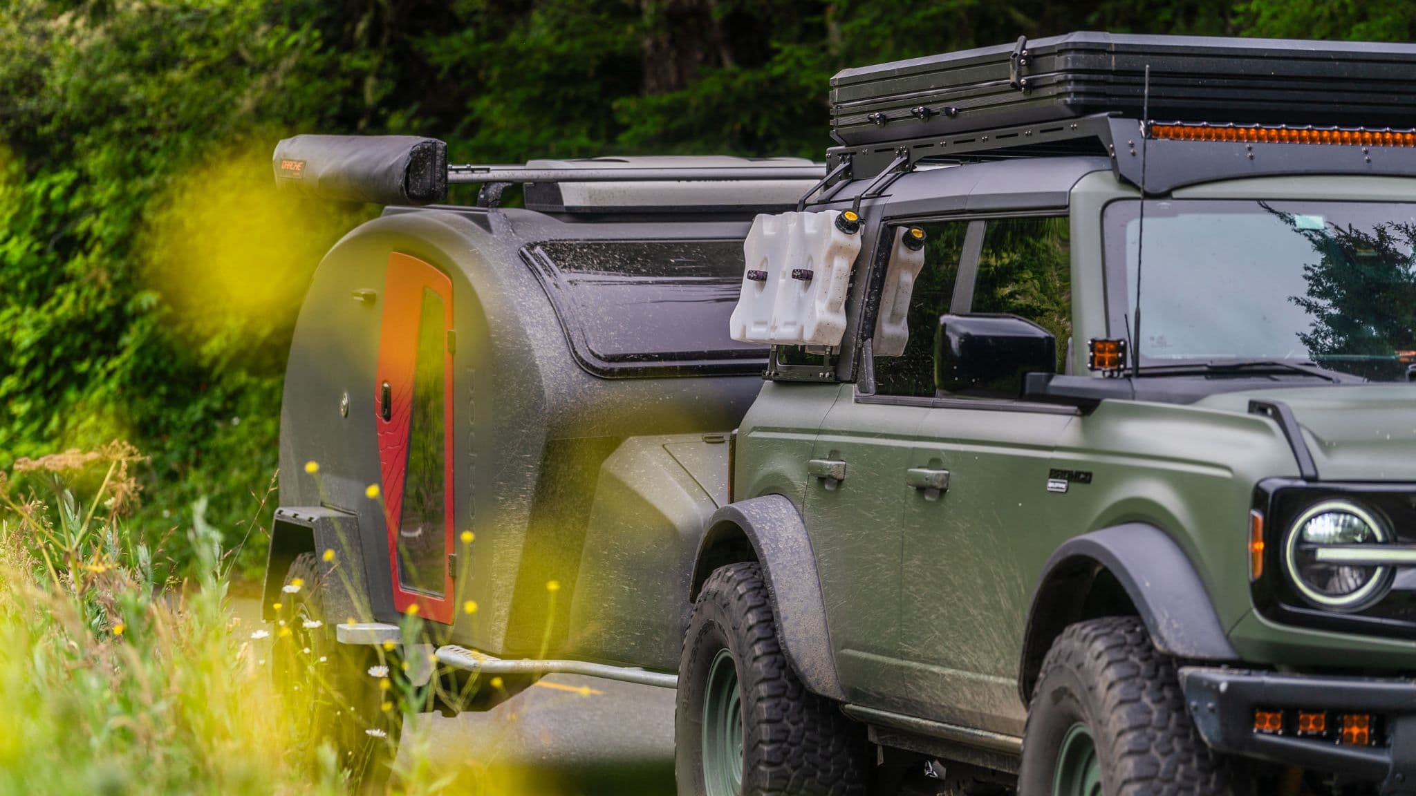 A close-up of an Escapod TOPO2 trailer hitched to a rugged off-road Bronco, showing its red-accented door and mounted water jugs against a backdrop of lush foliage.