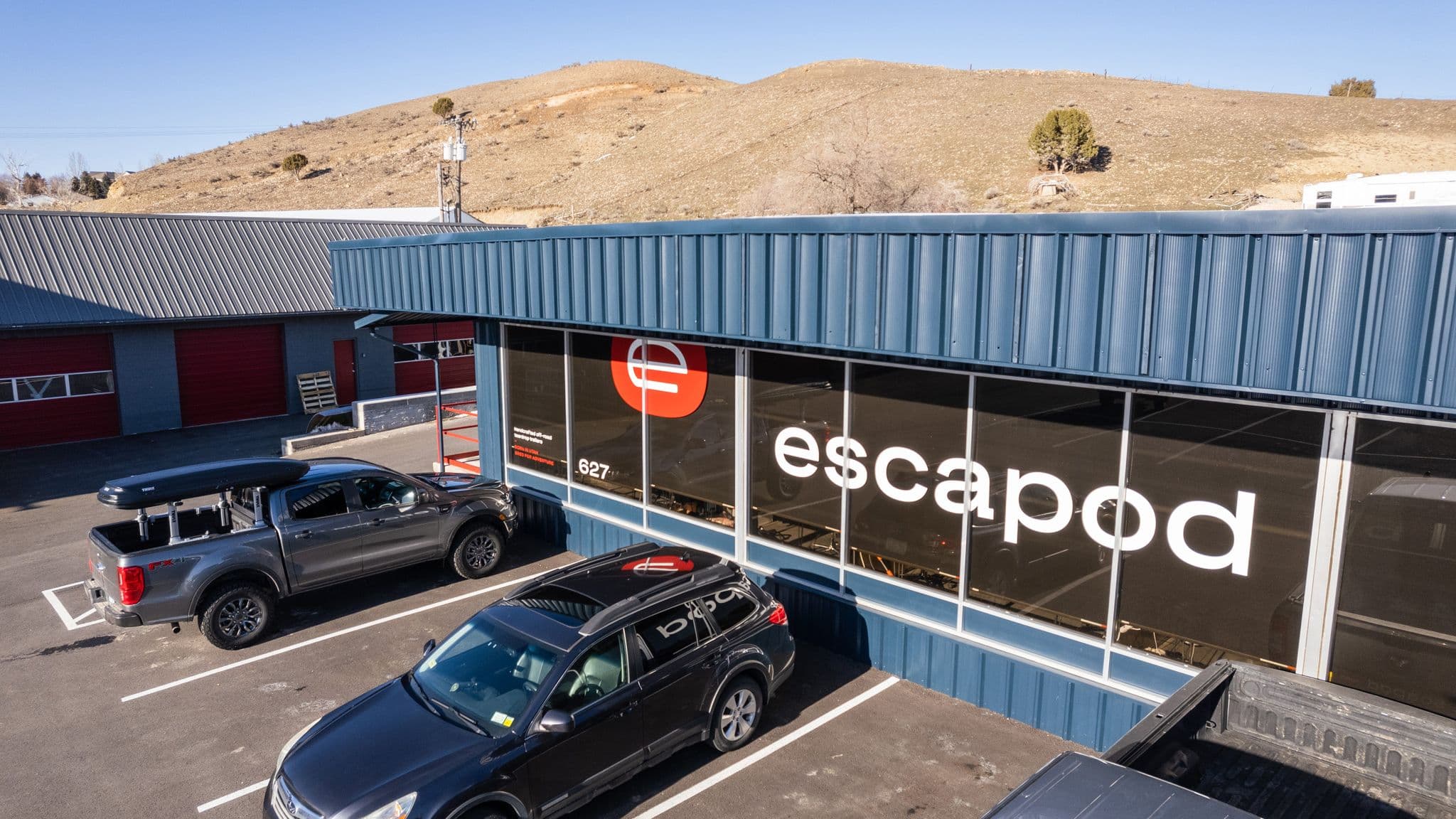 Aerial view of the Escapod facility showing the blue building with large "Escapod" branding, parking lot, and surrounding hilly landscape under a clear sky.