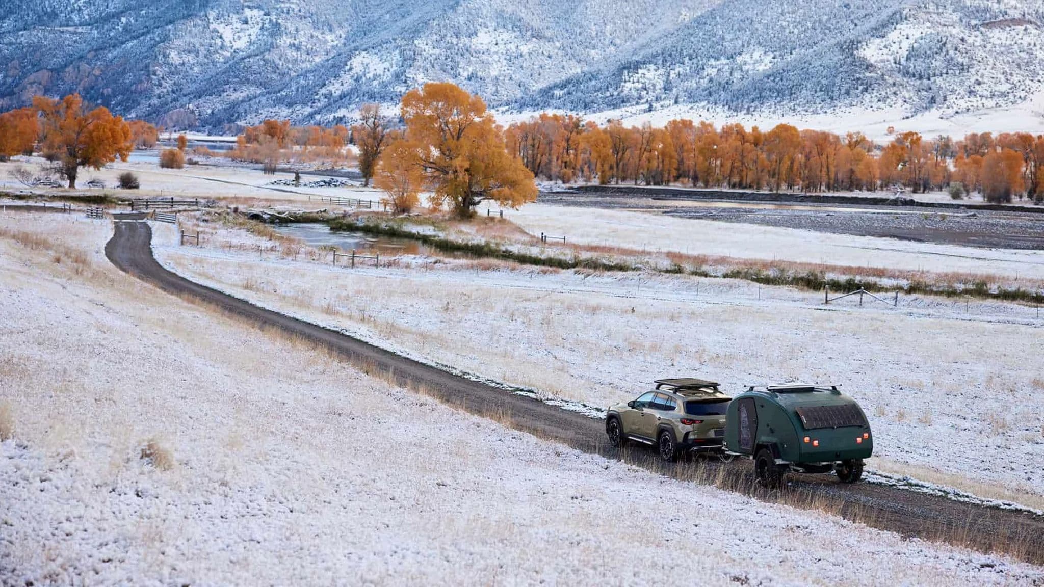 A Mazda CX-50 towing an Escapod TOPO2 trailer through a winding snow-covered valley, with orange autumn trees and towering mountains in the background.