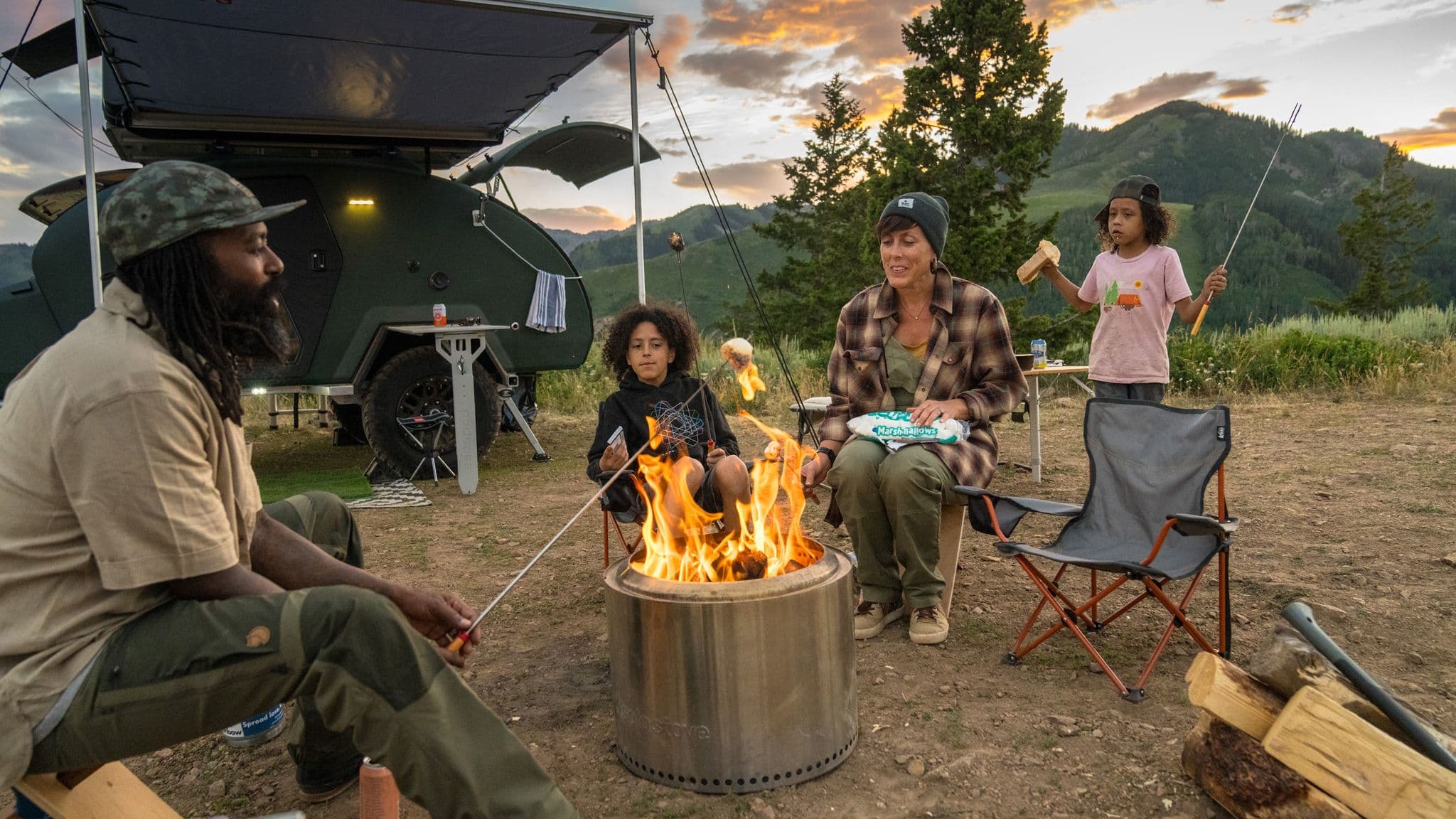 Family gathered around a campfire, roasting marshmallows at dusk, with an Escapod trailer set up under a mountain sunset.