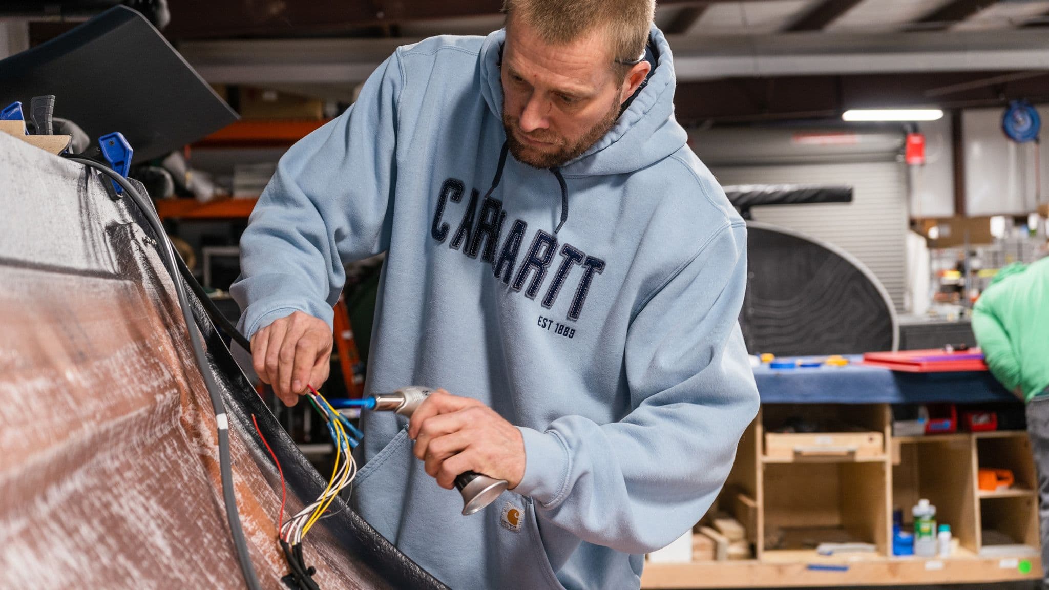 A technician in a blue Carhartt hoodie carefully connects electrical wiring inside an Escapod trailer assembly area.