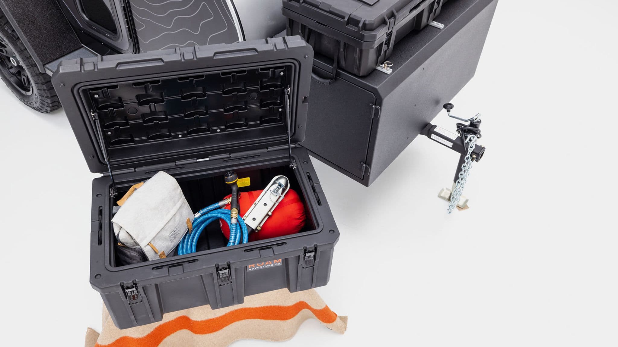 Open storage box at the front of the Escapod TOPO trailer, packed with camping gear, including a hose, a red sleeping bag, and a recovery tool.