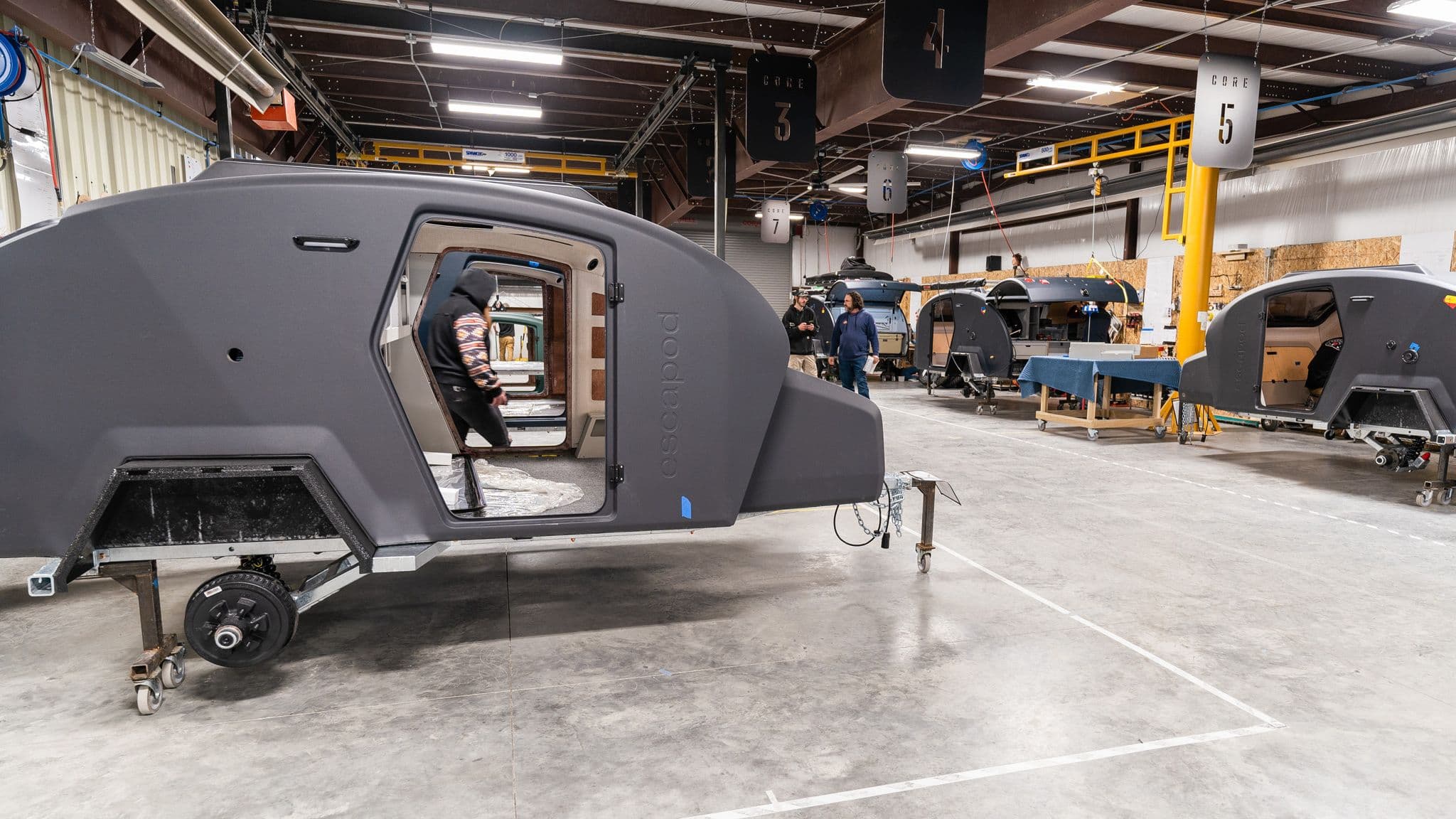 A row of Escapod TOPO2 trailers in different stages of assembly on the production floor, with workers handling various build tasks.