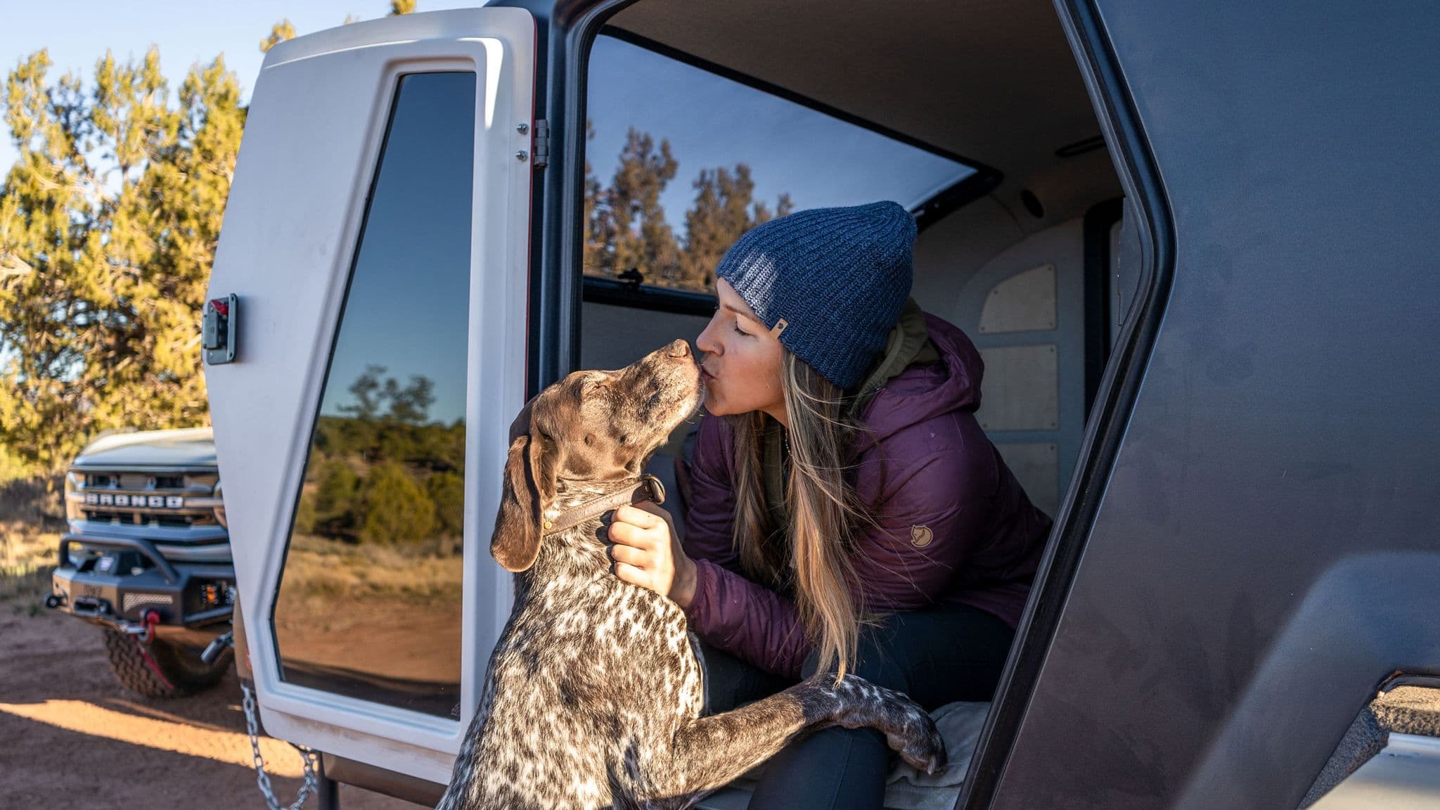 Woman in a beanie and maroon jacket kisses her brown and white dog while sitting in the doorway of an Escapod trailer, with a Bronco in the background.