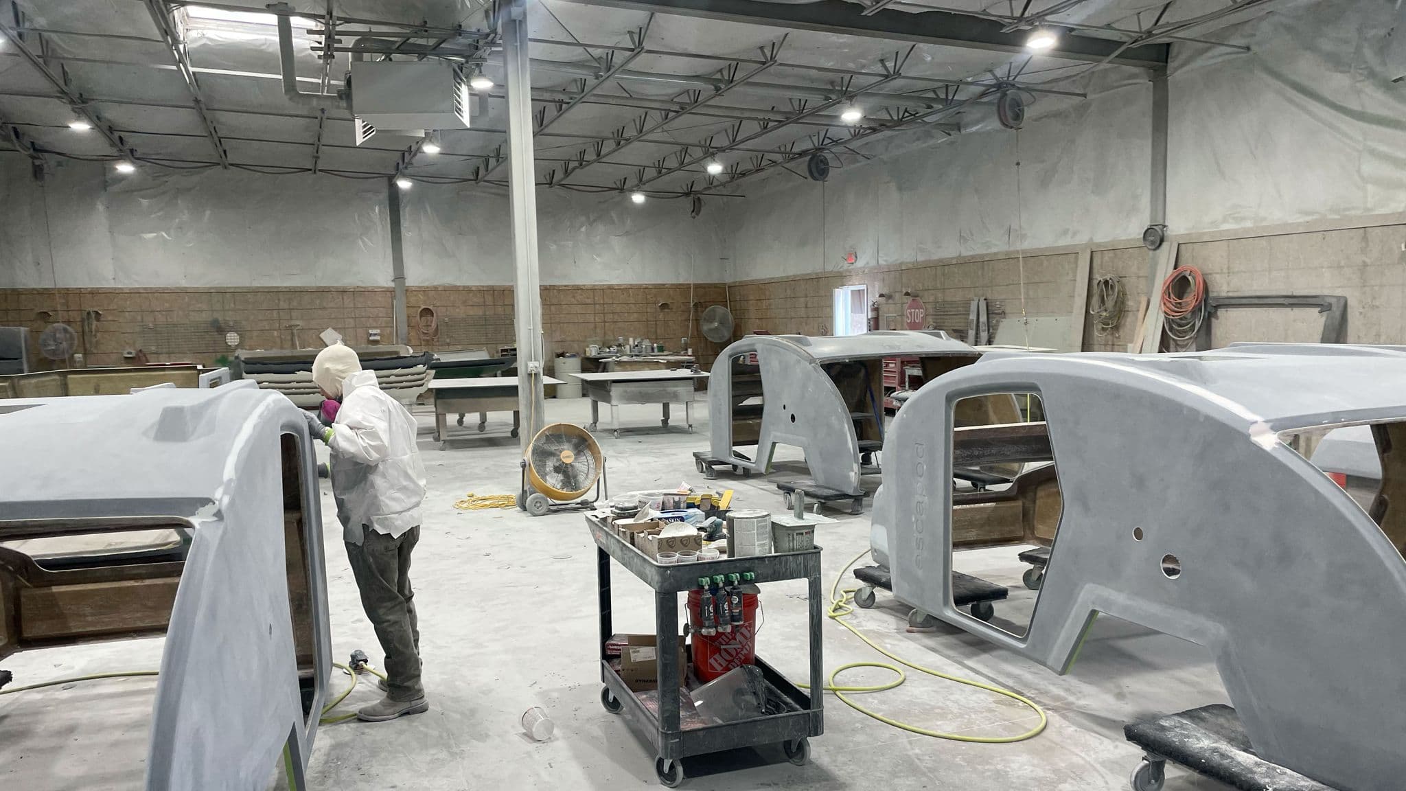 A wide view of Escapod’s factory floor, showing multiple teardrop trailer bodies in various stages of production, with a worker in protective gear sanding the surface.