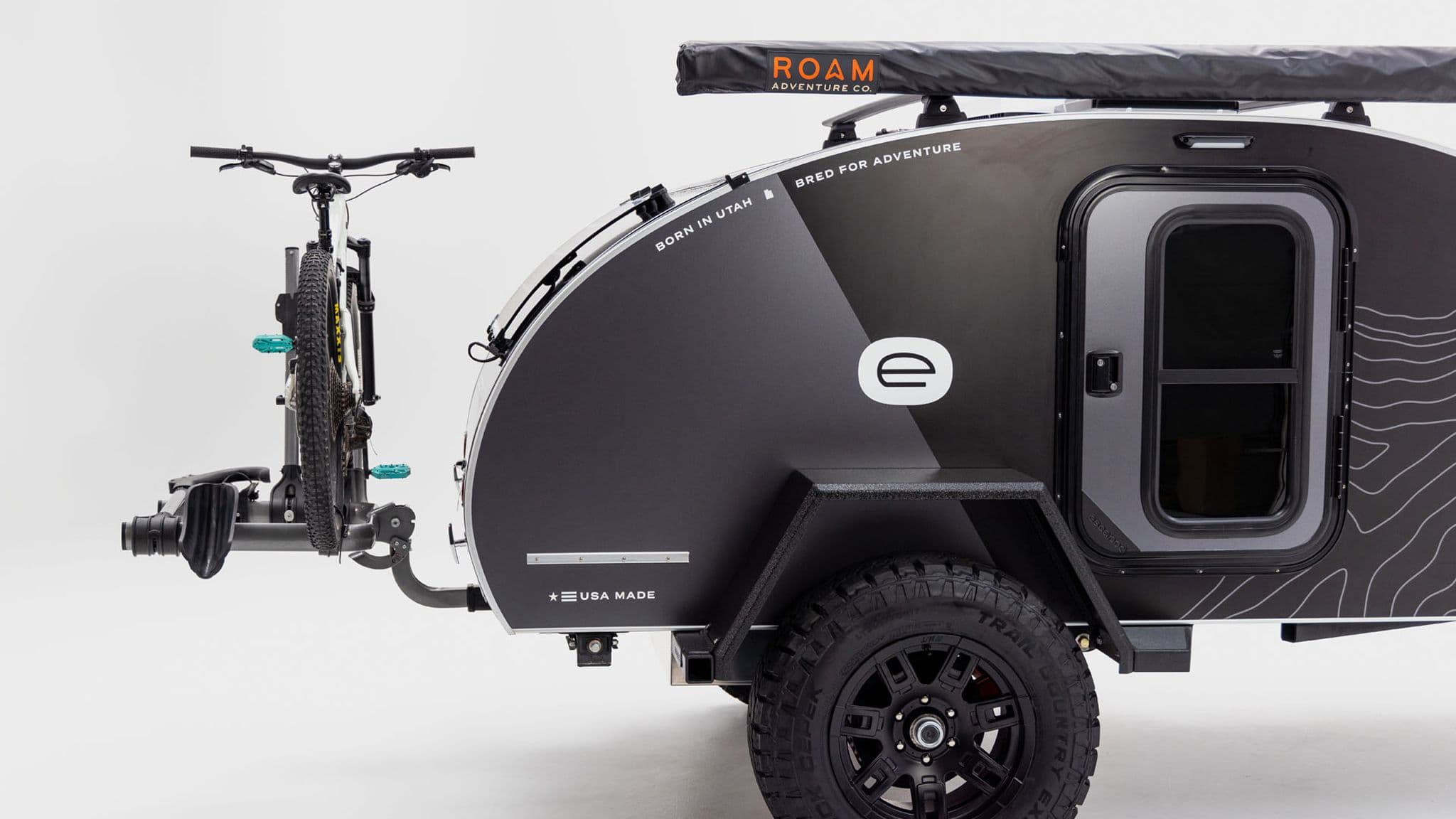 Side profile of an Escapod TOPO teardrop trailer with a mountain bike mounted on a front bike rack and a rooftop awning.