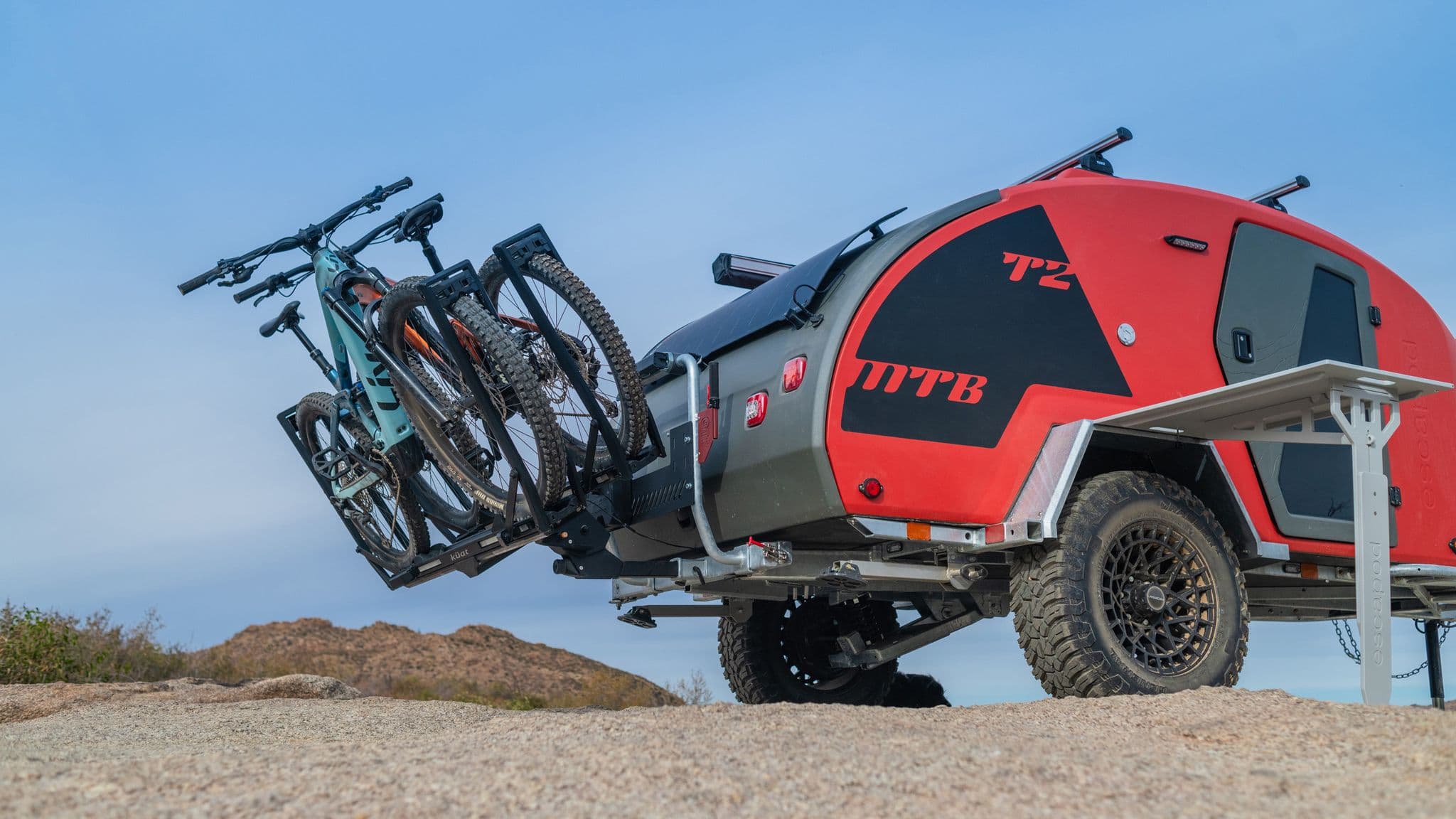 Two mountain bikes secured to the swing-away rear rack of an Escapod TOPO2 in lava orange, with the galley side table extended.