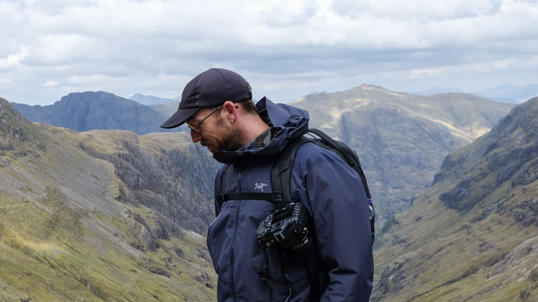 A man in a dark blue rain jacket and black cap hikes through a rugged mountain range, carrying a professional camera strapped to his chest.