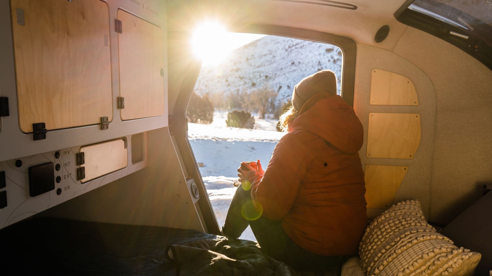 A camper enjoying a winter sunrise from inside an Escapod trailer, wrapped in a warm jacket while sipping coffee and looking out over a snow-covered landscape.