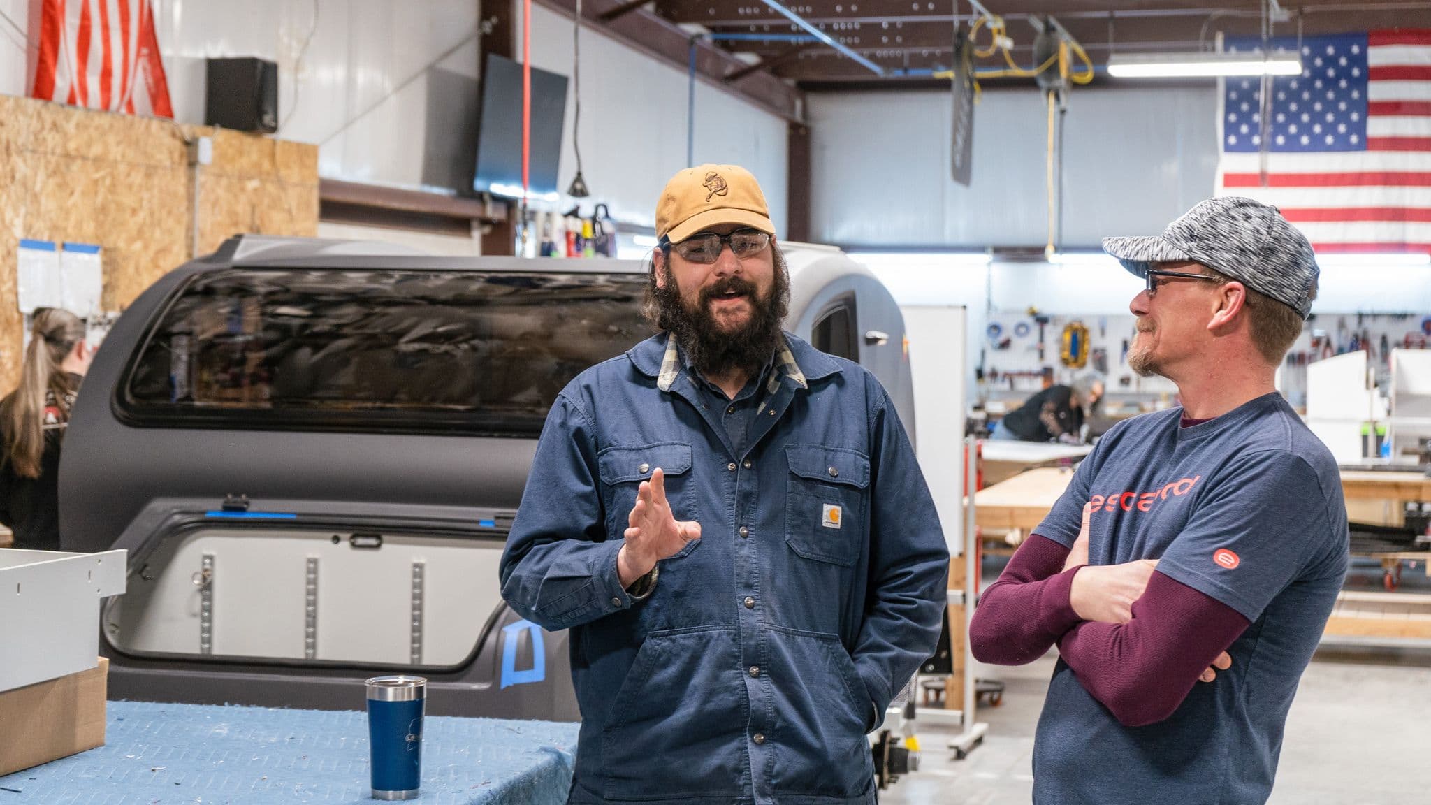 Two Escapod team members have a discussion in the factory, with one gesturing as he speaks. A partially assembled trailer and an American flag are visible in the background.