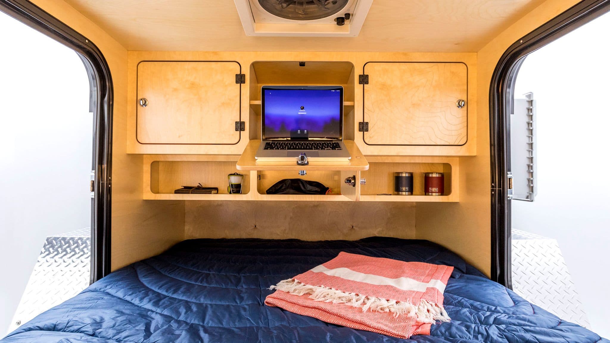 Interior of an Escapod TOPO teardrop trailer featuring a fold-out laptop desk, wooden storage cabinets, and a cozy sleeping area.