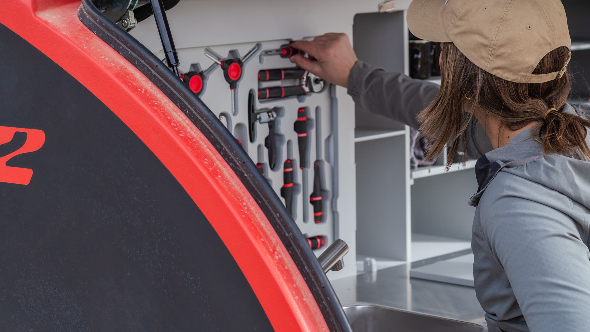 Woman selects screwdriver from tool panel in Escapod trailer galley with red TOPO2 MTB exterior visible.