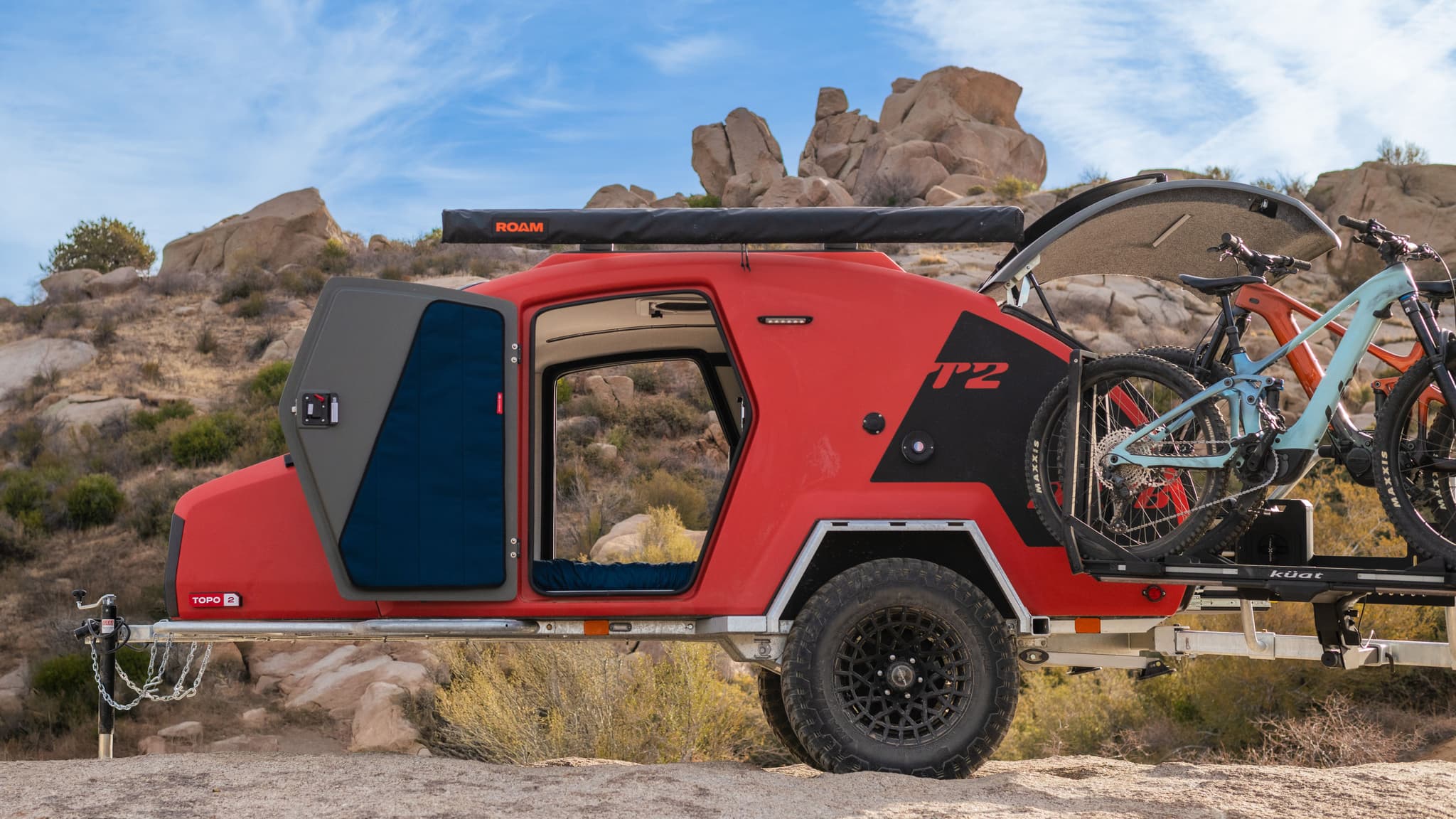 Side view of the TOPO2 MTB trailer with the door open and both bikes mounted on the rear rack, set against a rocky desert backdrop.