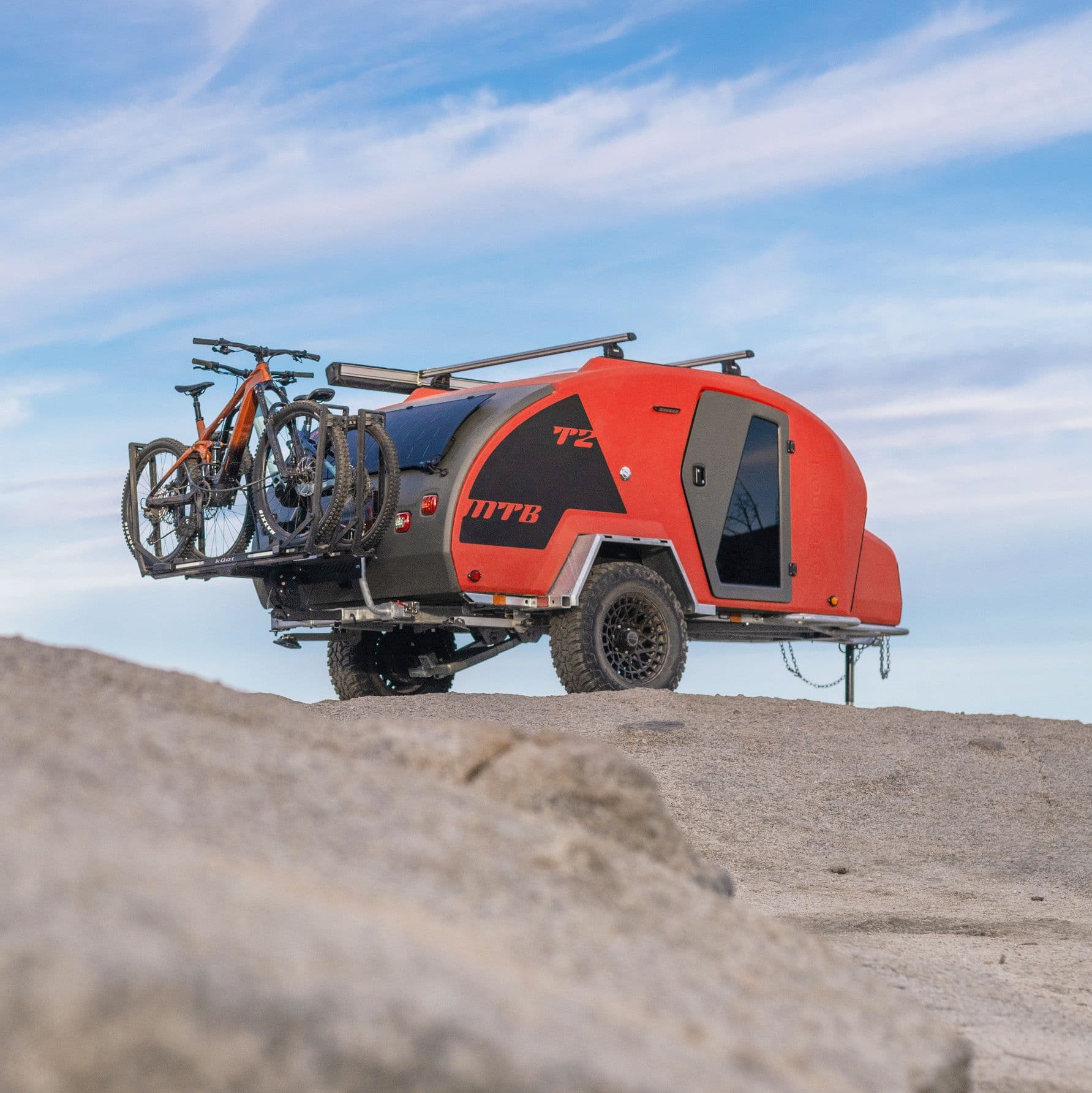 A Lava Orange TOPO2 MTB with two full suspension mountain bikes mounted on the rear, photographed on a rocky terrain from a low rear angle, with blue sky and wispy clouds.