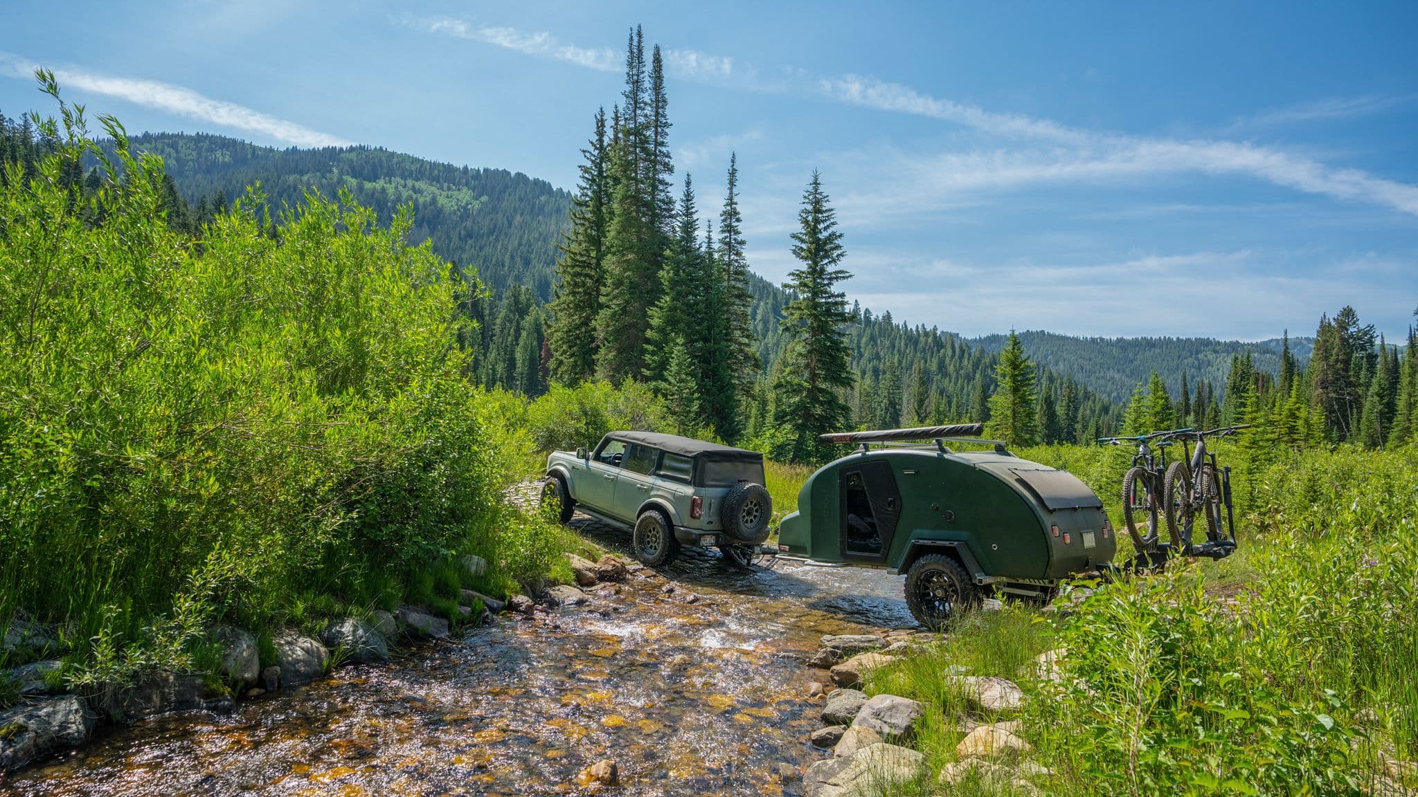 Ford Bronco towing an Escapod trailer across a shallow mountain stream, surrounded by lush greenery and towering evergreen trees.