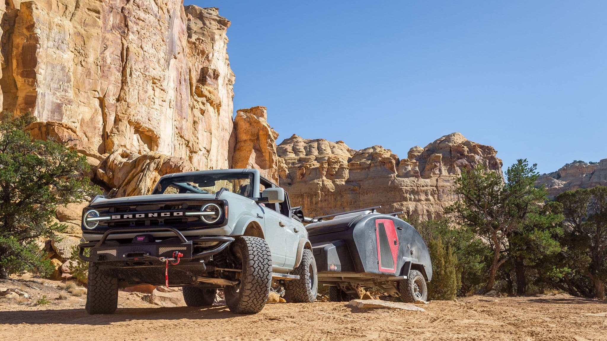 Ford Bronco towing an Escapod trailer in a stunning desert landscape with towering sandstone cliffs and bright blue sky, built for adventure.