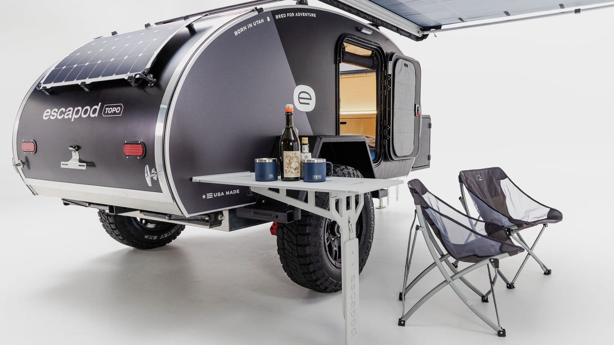 Rear view of the Escapod TOPO trailer in a studio setting, with a fold-out side table, camping chairs, and a bottle of wine on the table.