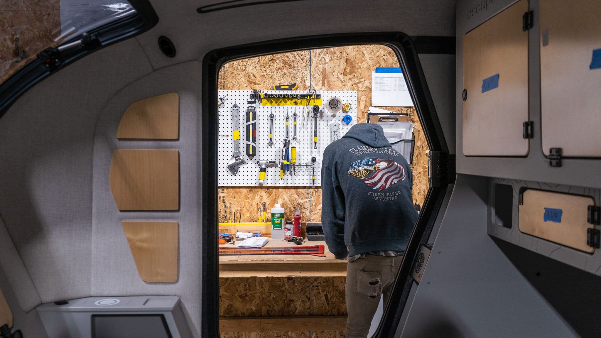 A view from inside an Escapod trailer looking out at a workstation, where a worker in a hoodie stands in front of a pegboard filled with tools.
