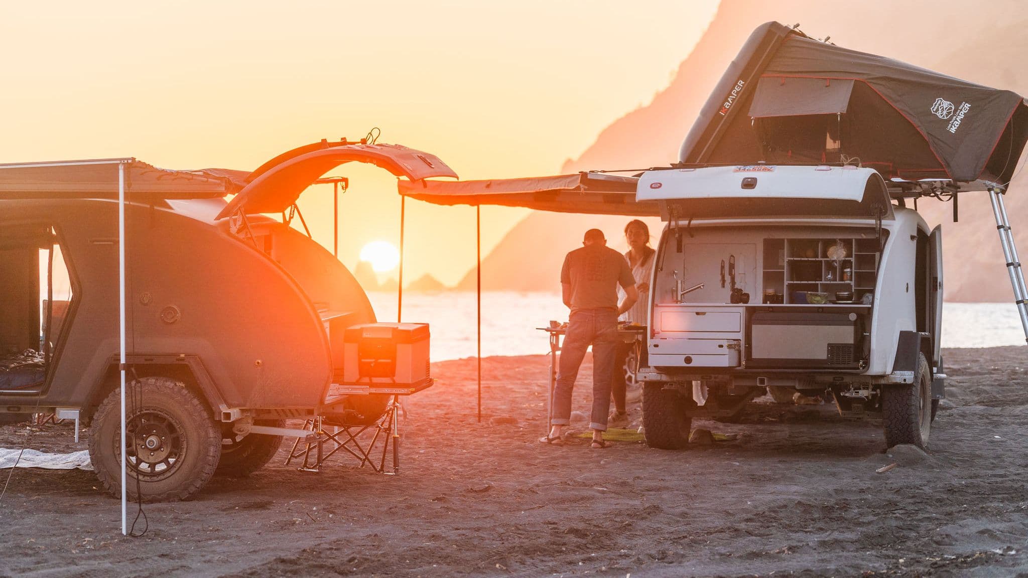 Two Escapod trailers parked on a beach at sunset, with a couple preparing a meal under the glow of the golden sky.