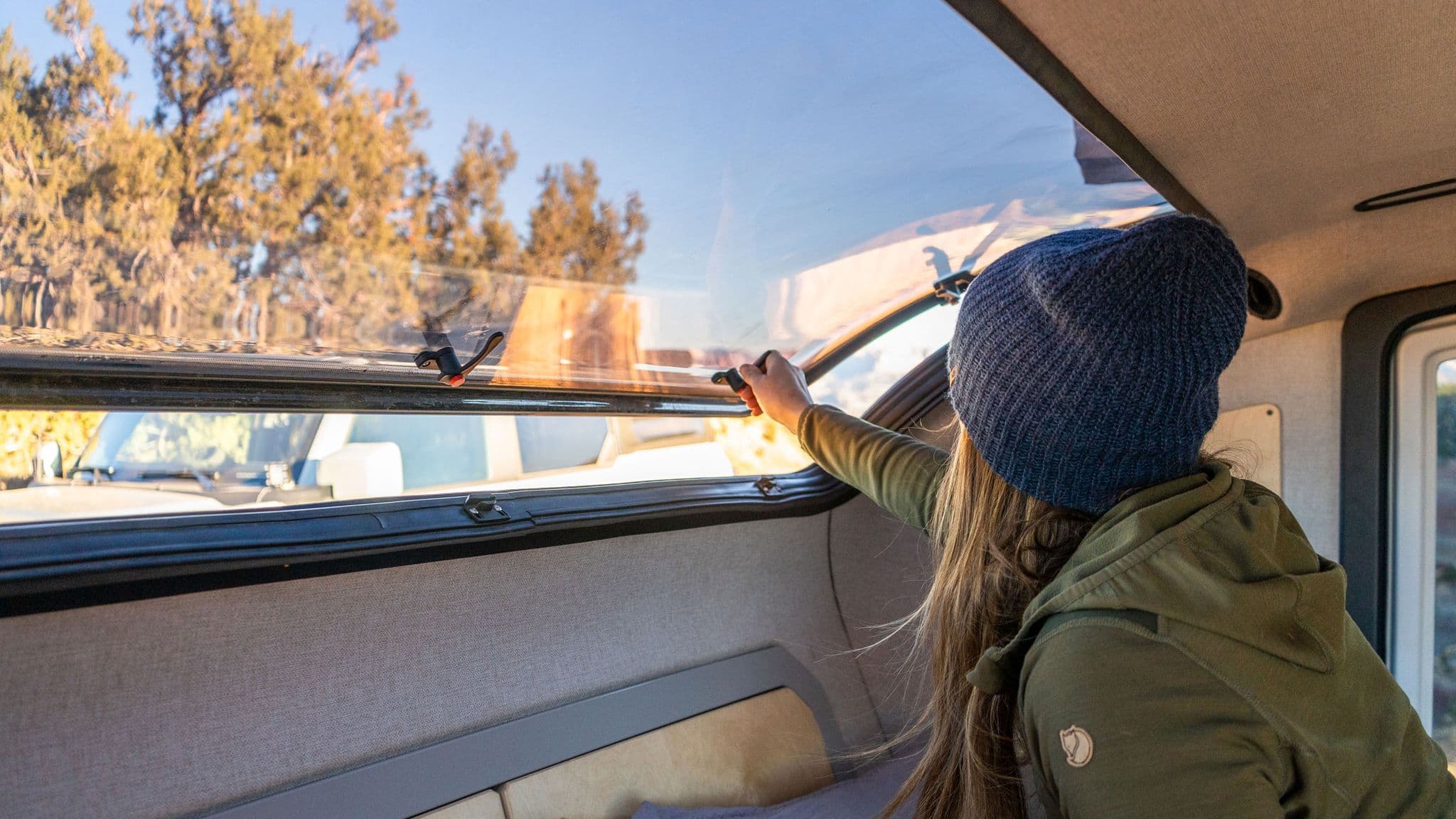 Camper adjusts the large acrylic skylight in an Escapod trailer, allowing natural light and ventilation while parked in a scenic forest location.