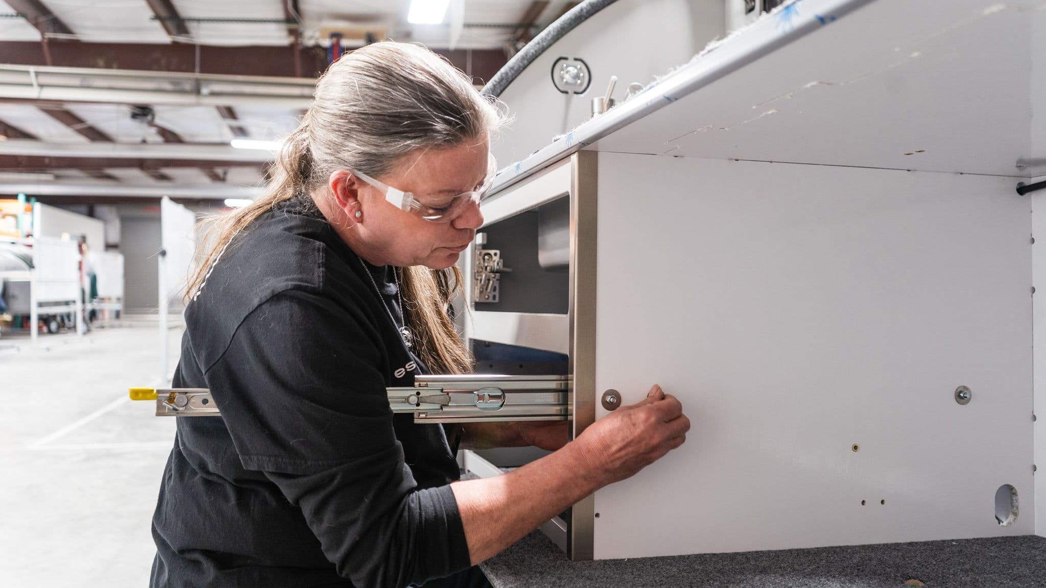 A factory worker installs cabinetry inside an Escapod trailer, carefully securing a panel while wearing safety glasses.