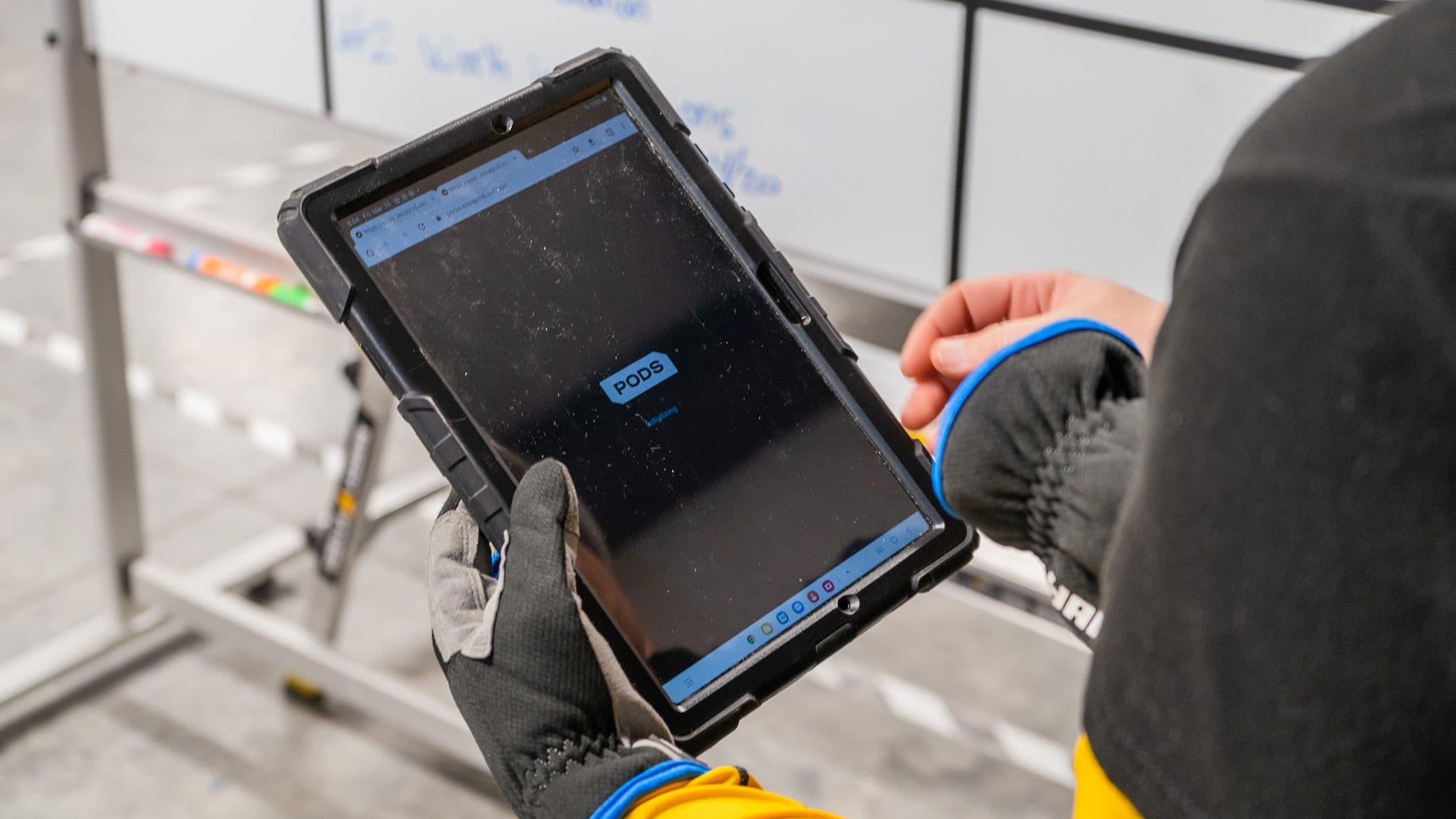 A worker in gloves holds a rugged tablet displaying "PODS," Escapod's proprietary quality control software, in front of a whiteboard with handwritten notes in the factory.