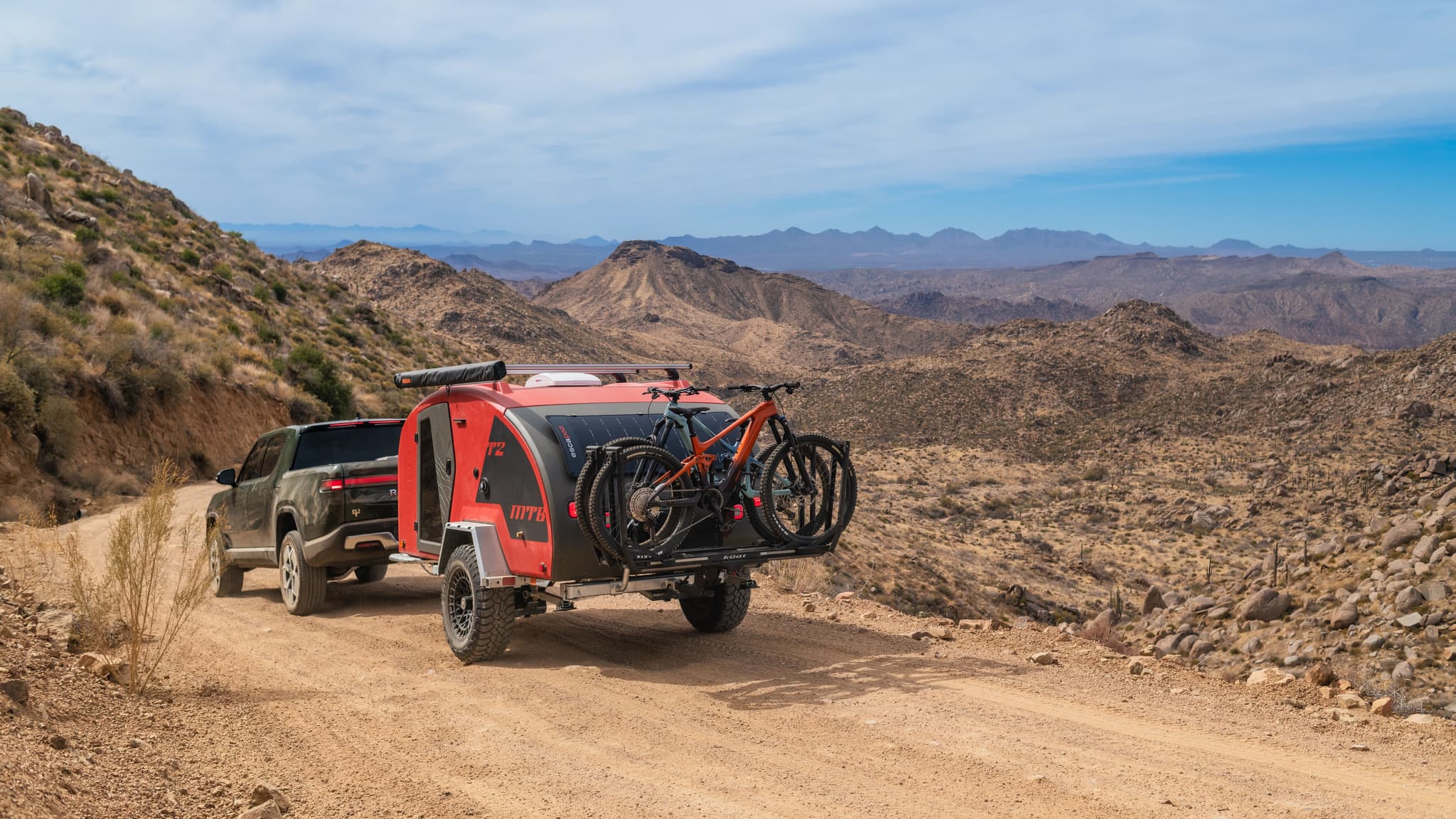 Wide landscape shot of a Rivian R1T towing the Escapod TOPO2 MTB trailer, fully loaded with mountain bikes, on a winding dirt trail in the desert.