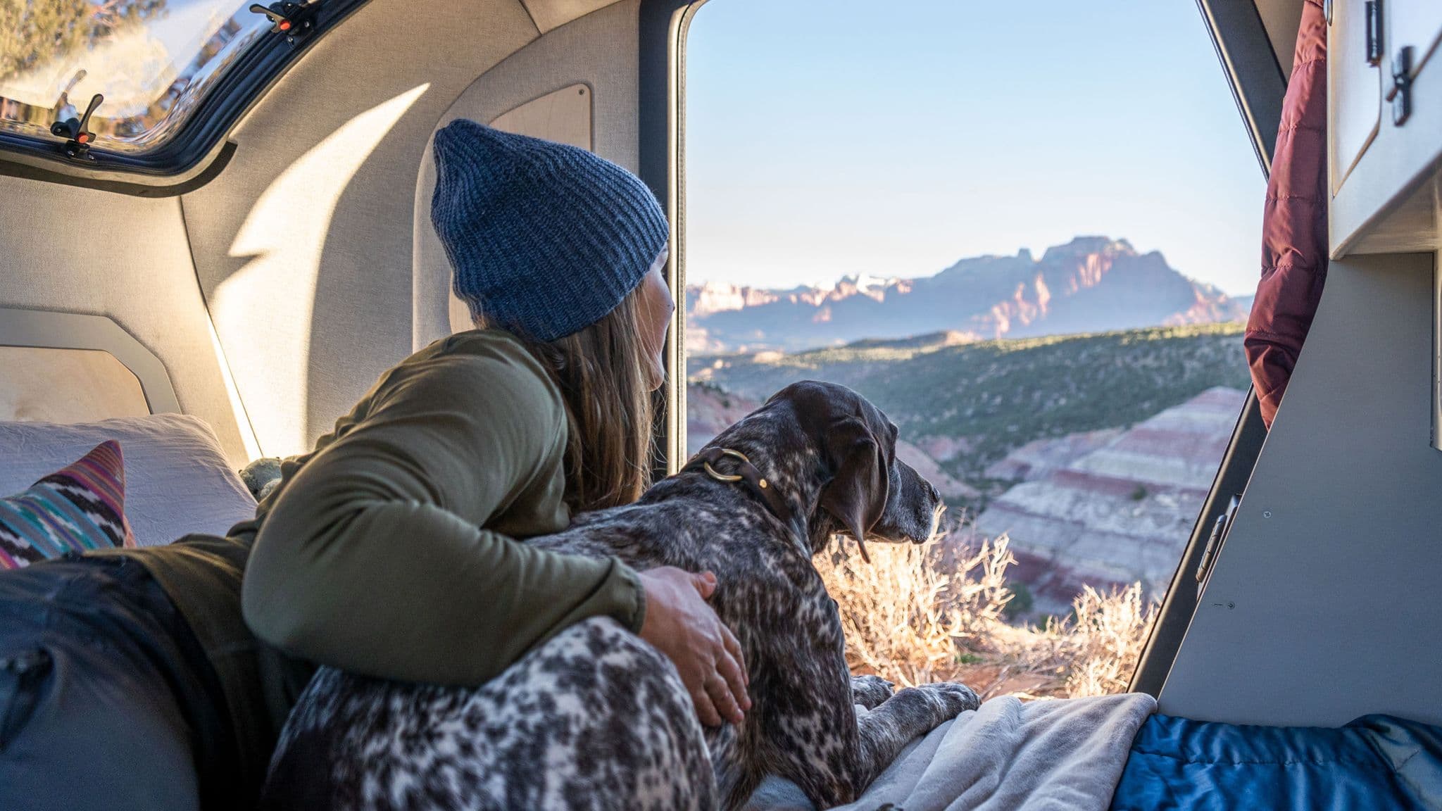 Woman in a beanie and green jacket lounges inside an Escapod trailer with her dog, enjoying a scenic mountain view through the open side door.