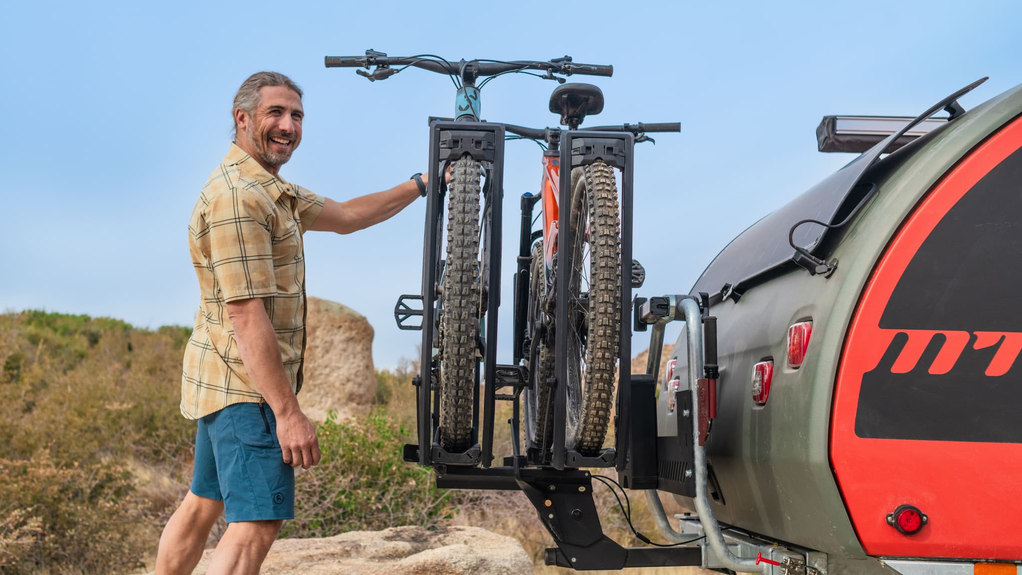 Smiling man in plaid shirt stands next to Escapod trailer, adjusting bikes mounted on rear bike rack.