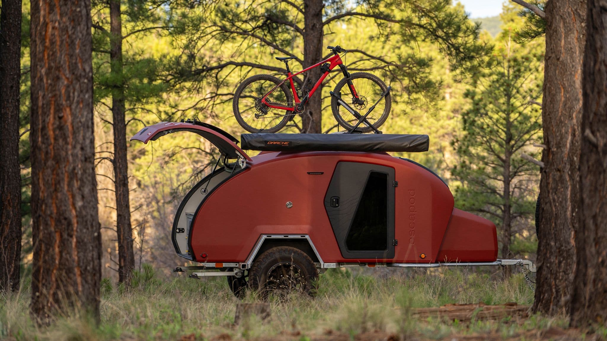 Escapod TOPO2 trailer parked among towering pine trees, equipped with a rooftop bike rack and awning, ready for off-grid adventure in the wild.