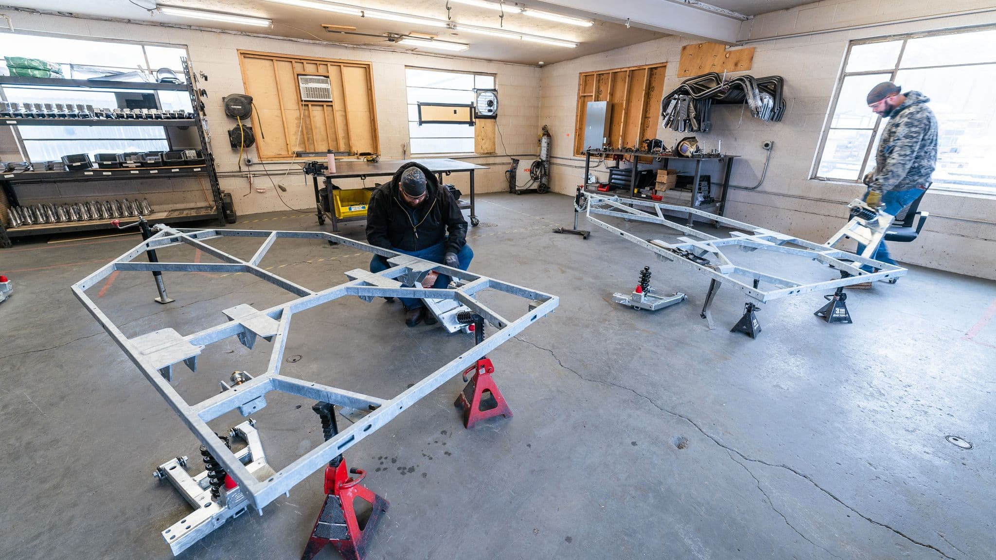 Two workers in a workshop assembling galvanized steel trailer chassis on red jack stands, surrounded by tools and equipment.