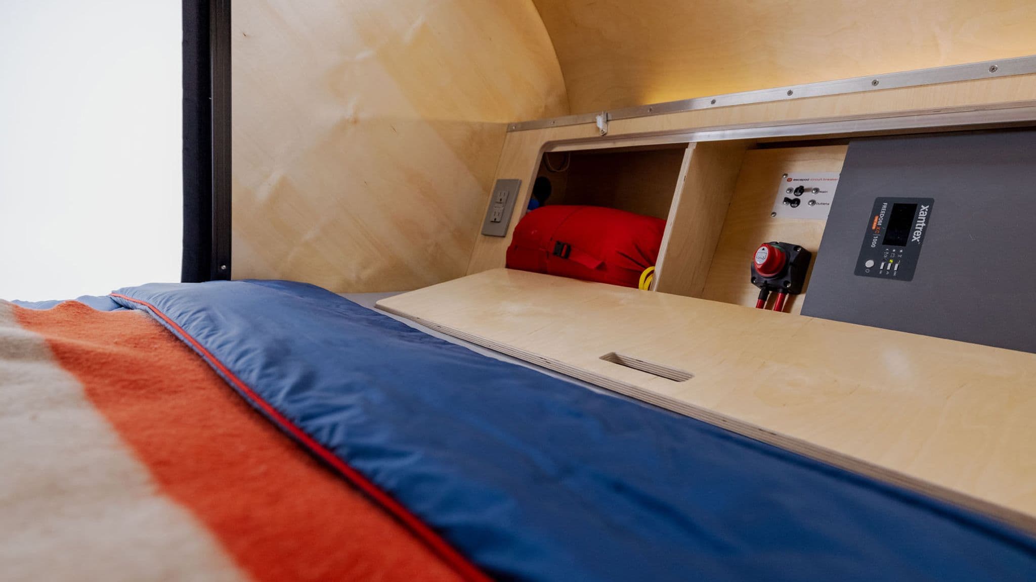 Interior sleeping area of the Escapod TOPO trailer, featuring a wooden storage shelf, control panel, and a cozy sleeping setup.