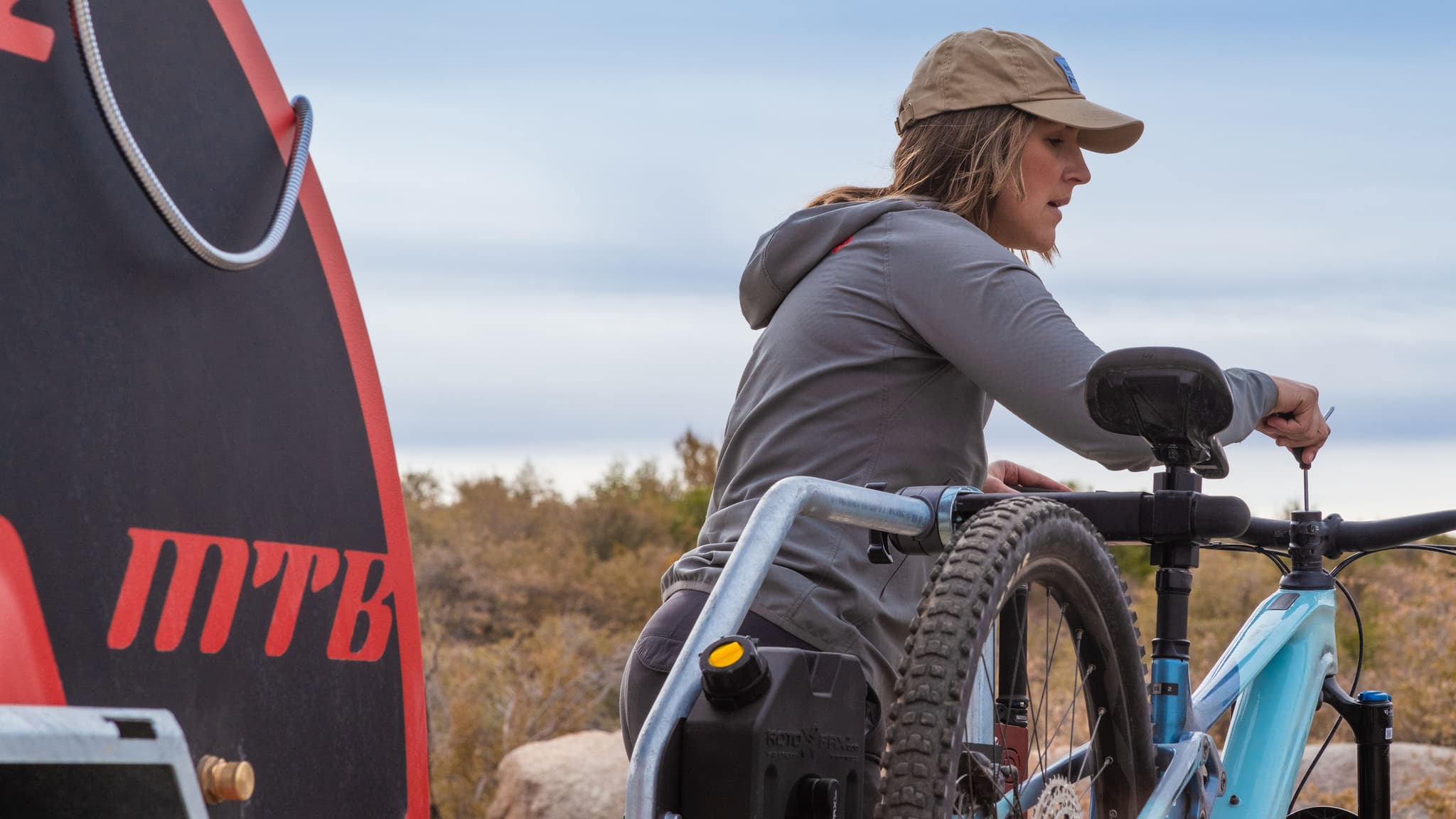 Woman uses a tool on handlebars of blue mountain bike secured to Escapod MTB trailer in desert.