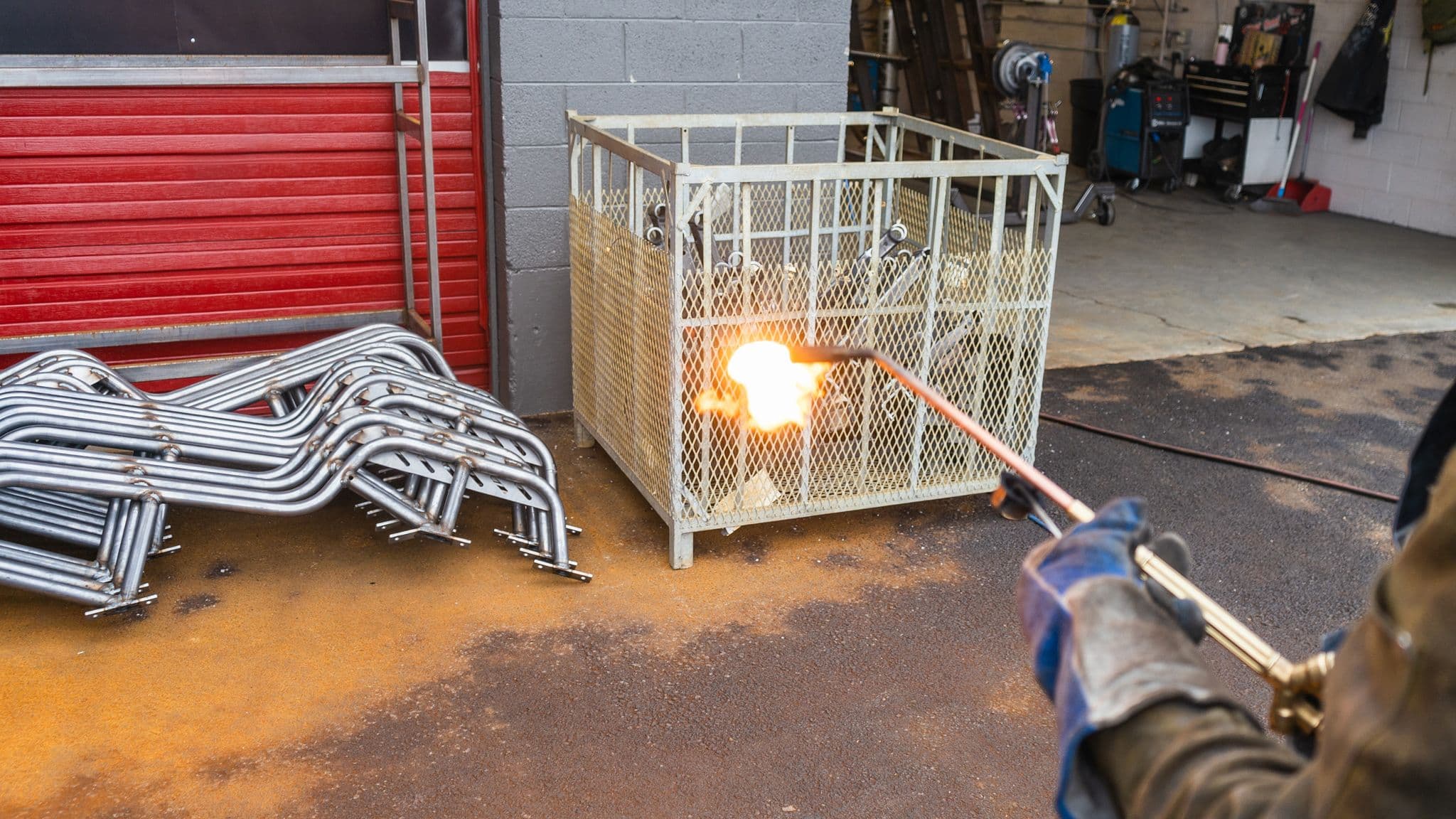 A factory worker uses a propane torch to heat metal components outside the Escapod facility, with flames igniting in front of a metal storage bin and red garage doors.