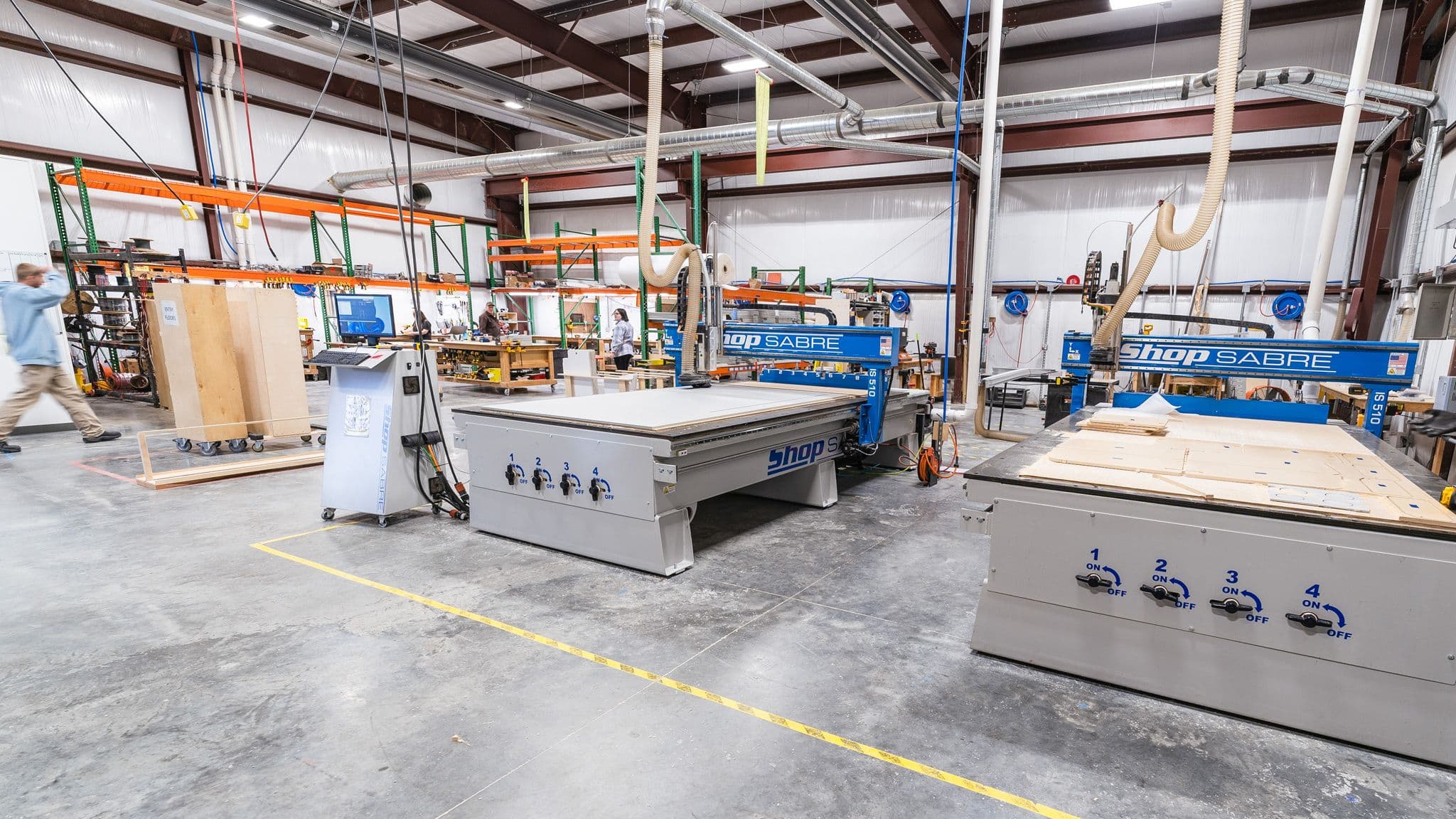 A spacious manufacturing facility with multiple large ShopSabre CNC machines, workstations, and employees working on various production tasks.