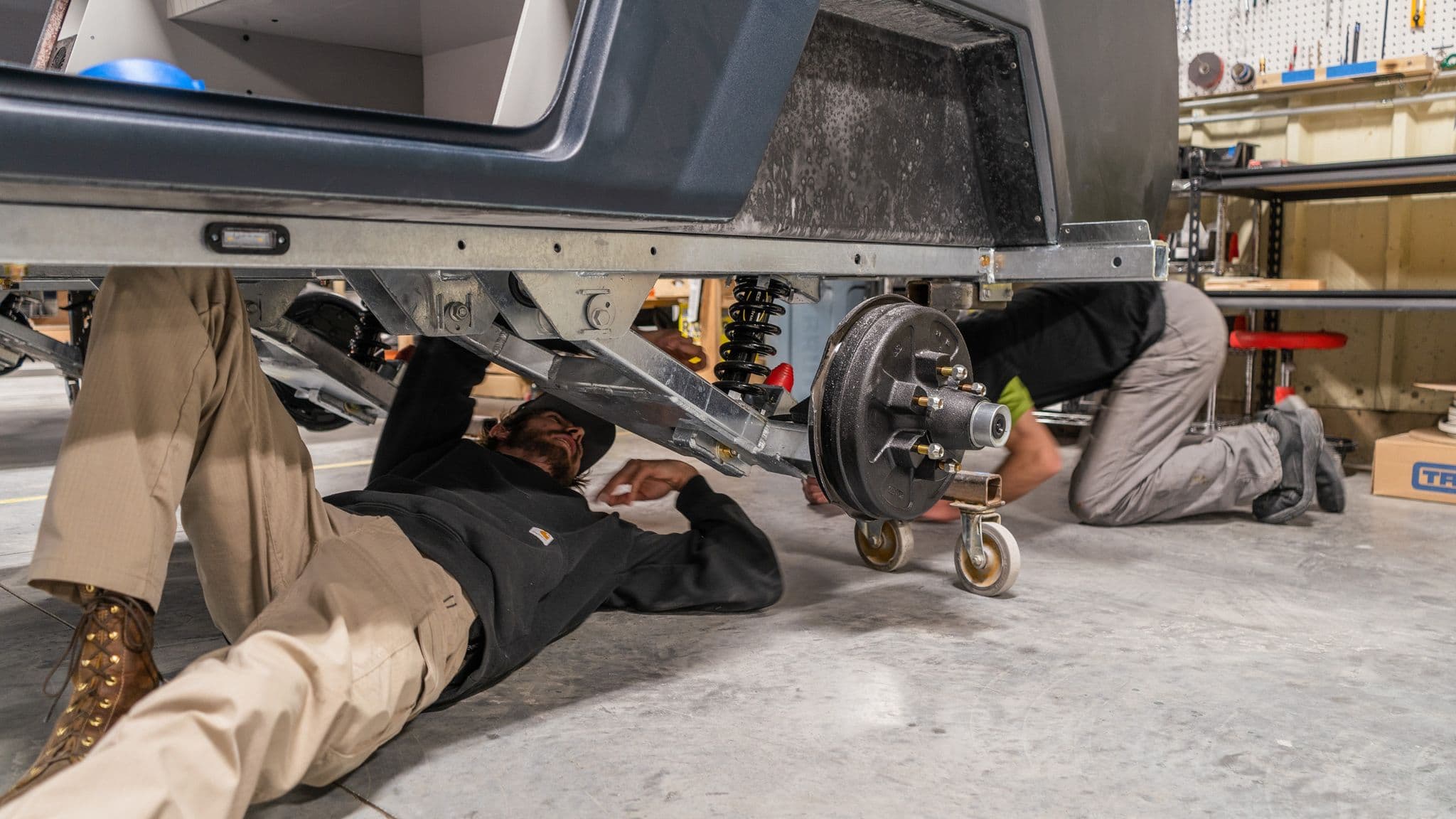 Two workers lay on their backs underneath an Escapod trailer, inspecting and assembling the suspension system in the factory.
