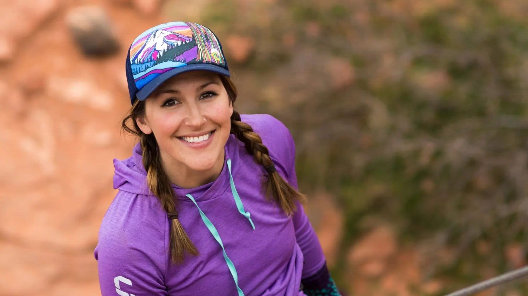 A woman in a vibrant purple hoodie and a colorful cap with mountain artwork smiles warmly, standing against a desert landscape with rocky terrain.