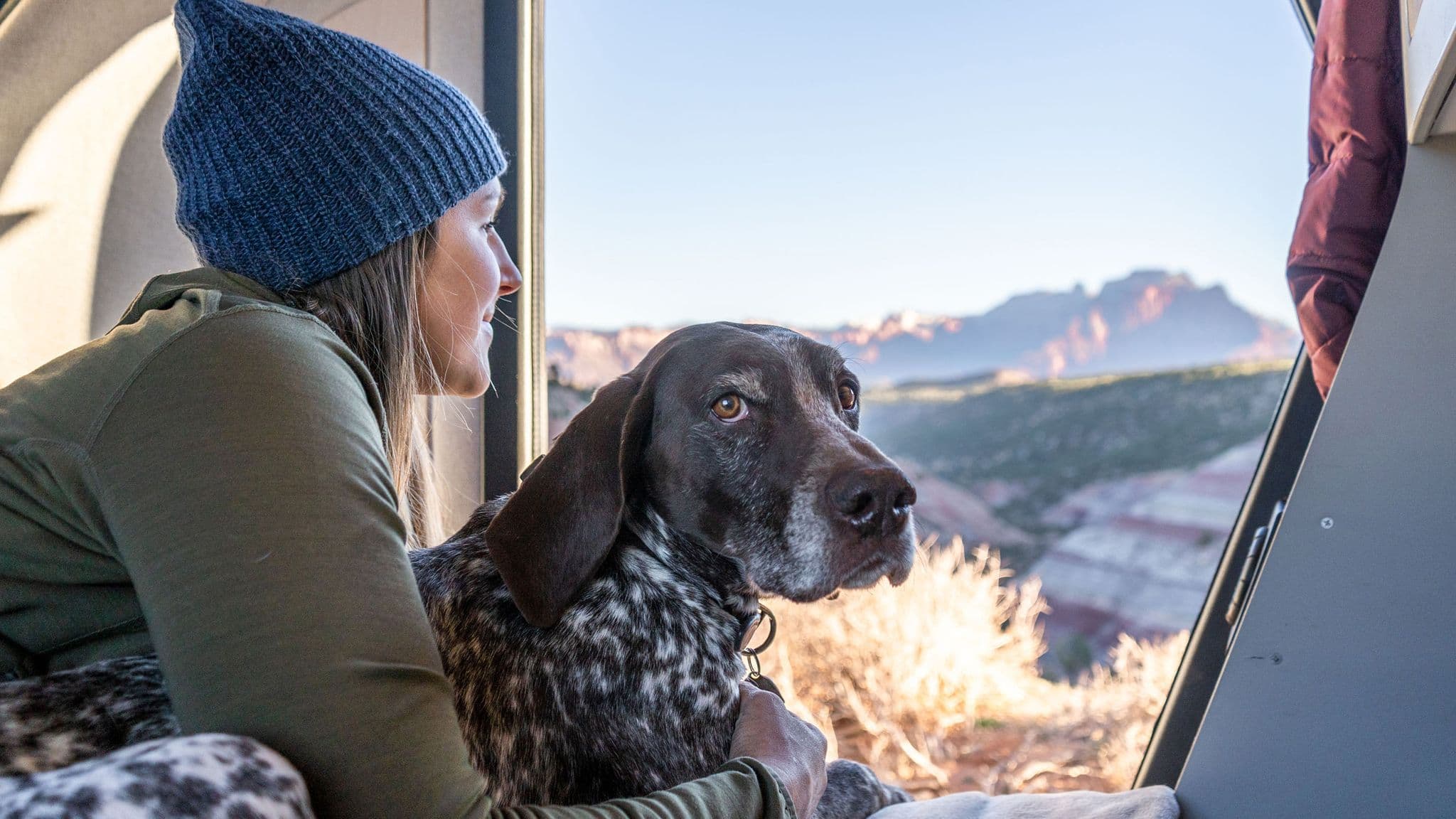 Woman and her dog rest inside an Escapod trailer, gazing out at Utah's colorful desert ridges, bathed in golden morning light.