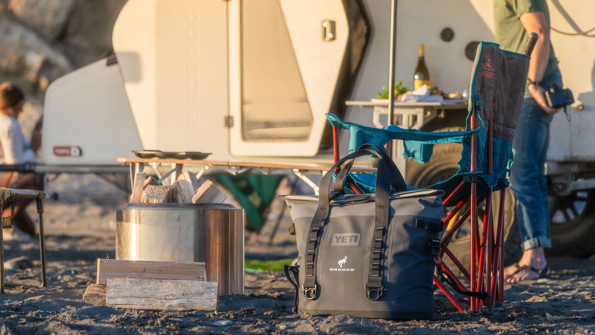 A Yeti cooler, camp chairs, and firewood set up near an Escapod trailer at a campsite on a sandy beach, with warm sunlight highlighting the outdoor setup.