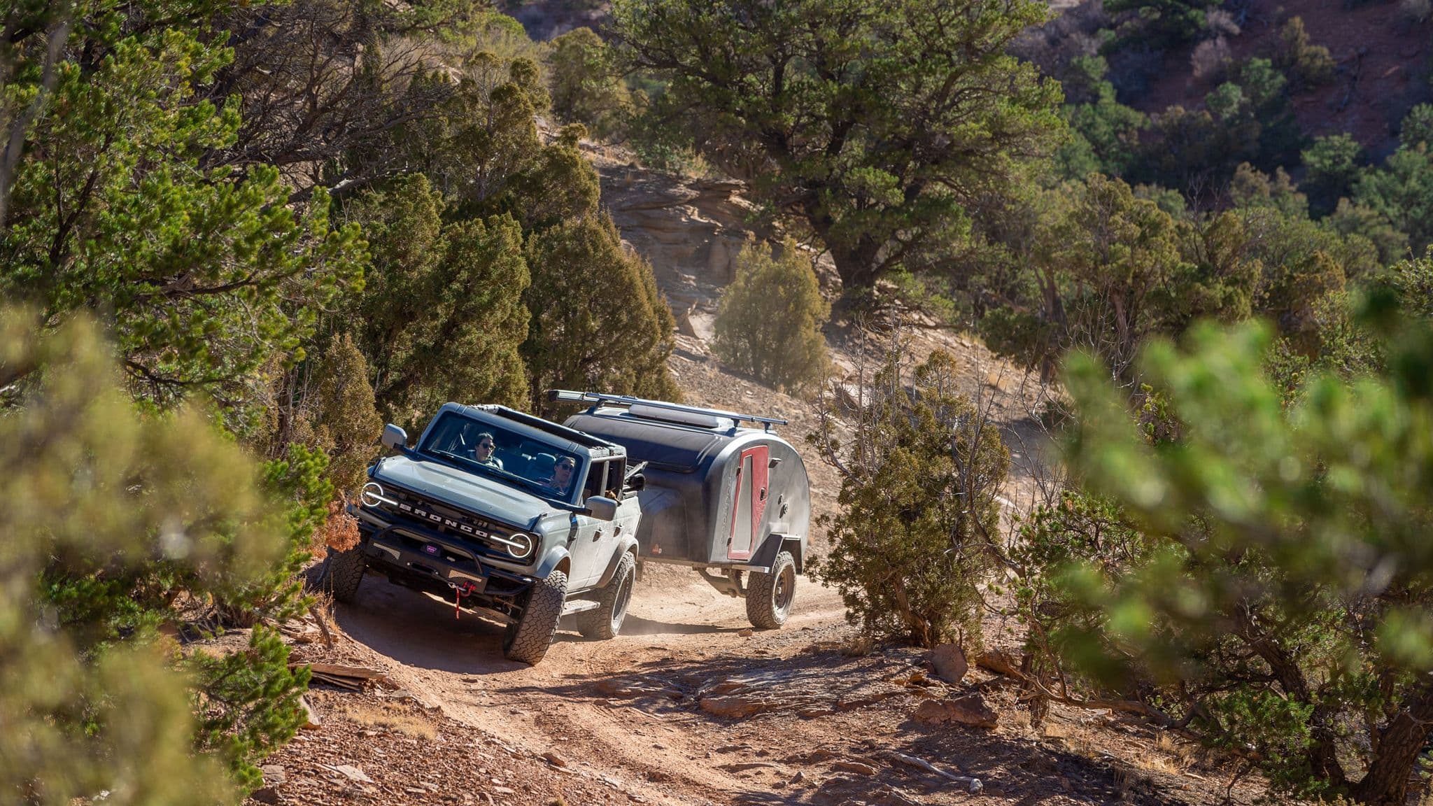 Ford Bronco towing an Escapod trailer through a winding dirt path in a high-desert setting.