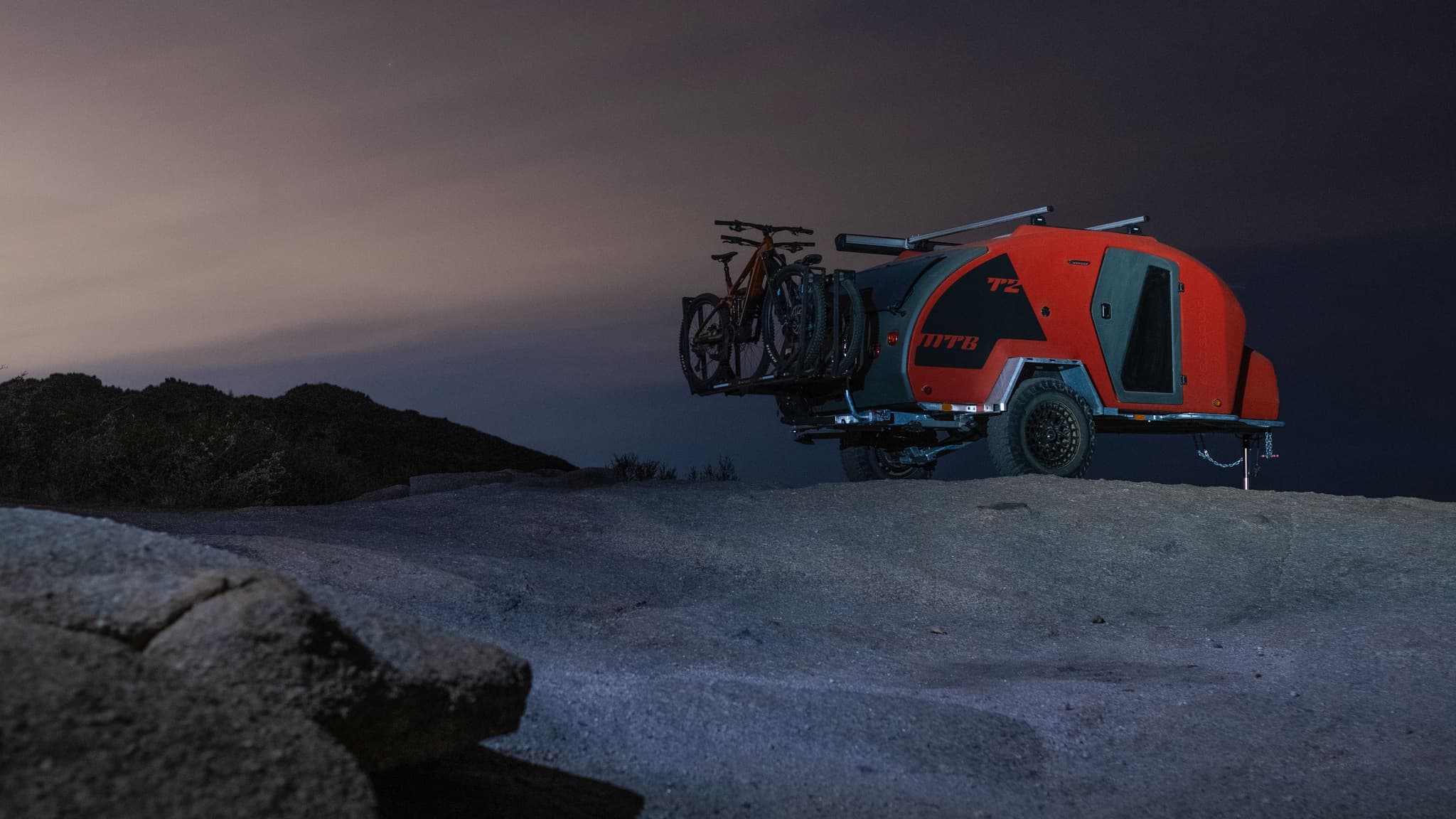 Red TOPO2 MTB trailer parked on rocky terrain at night, lit dramatically under a dark sky with mountain bikes mounted on the rear.