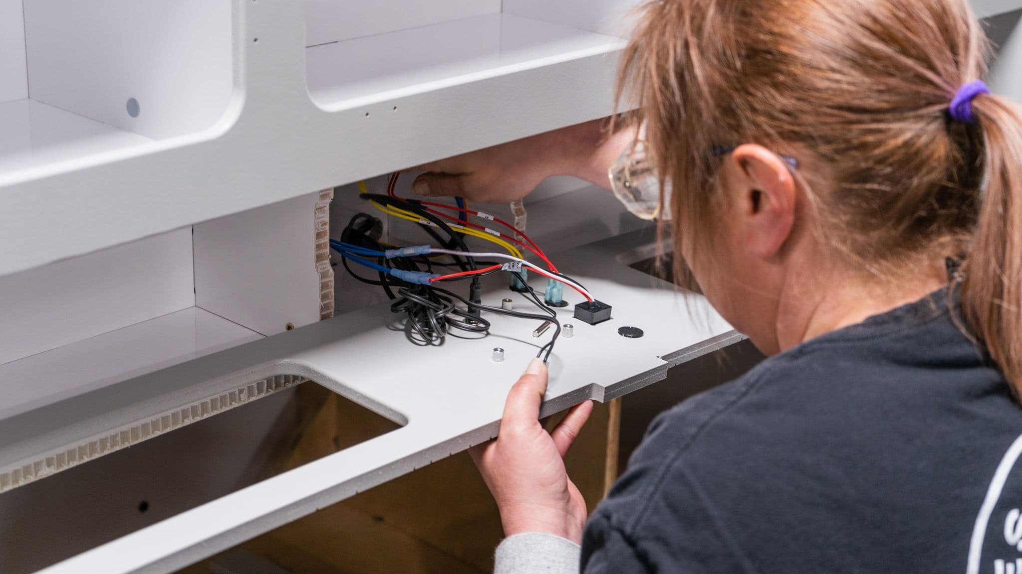 A female technician with safety glasses installs electrical wiring inside a white interior panel of an Escapod trailer.