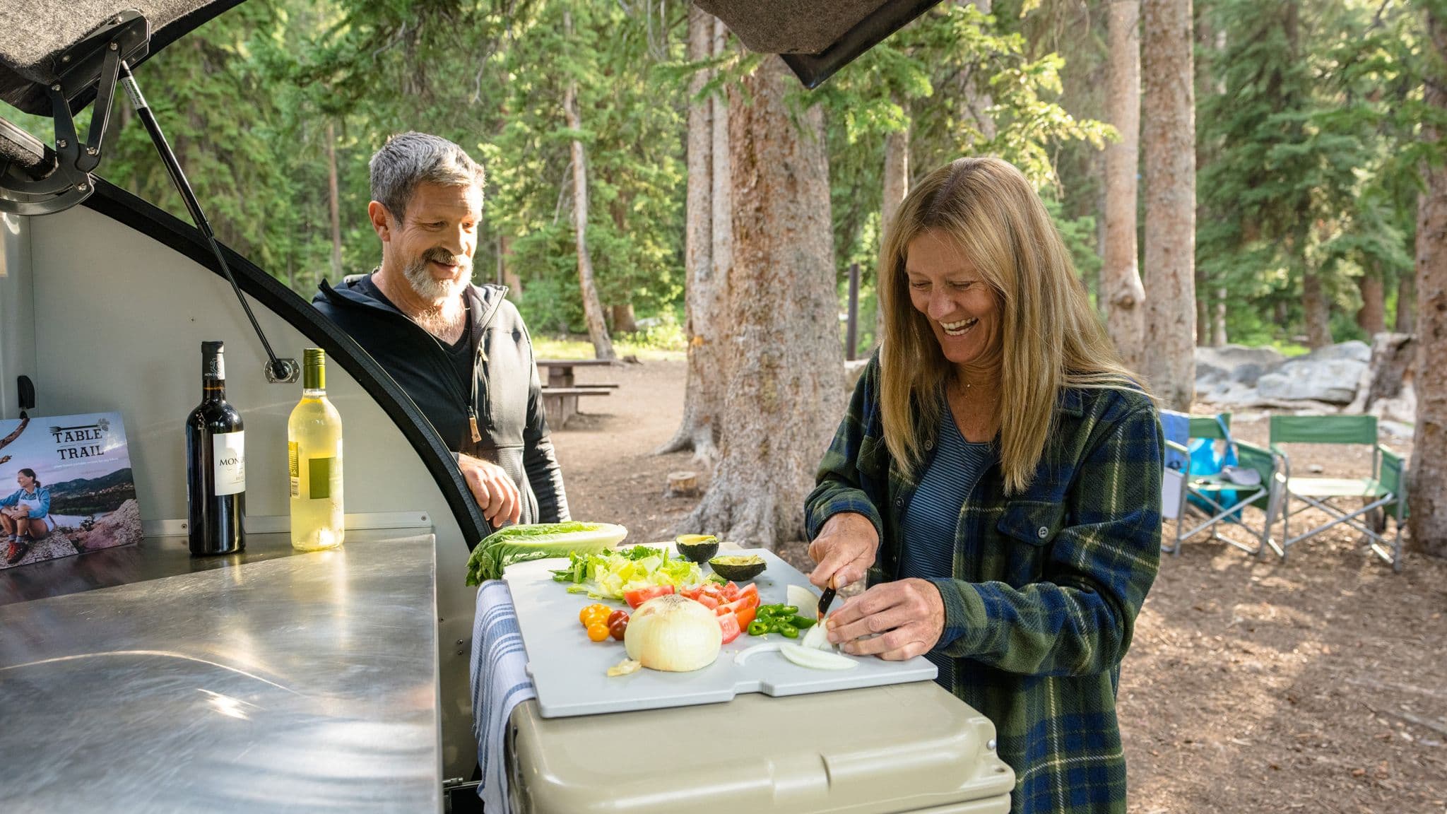 Couple preparing fresh vegetables in the galley kitchen of an Escapod trailer, surrounded by a lush forest campsite setting.